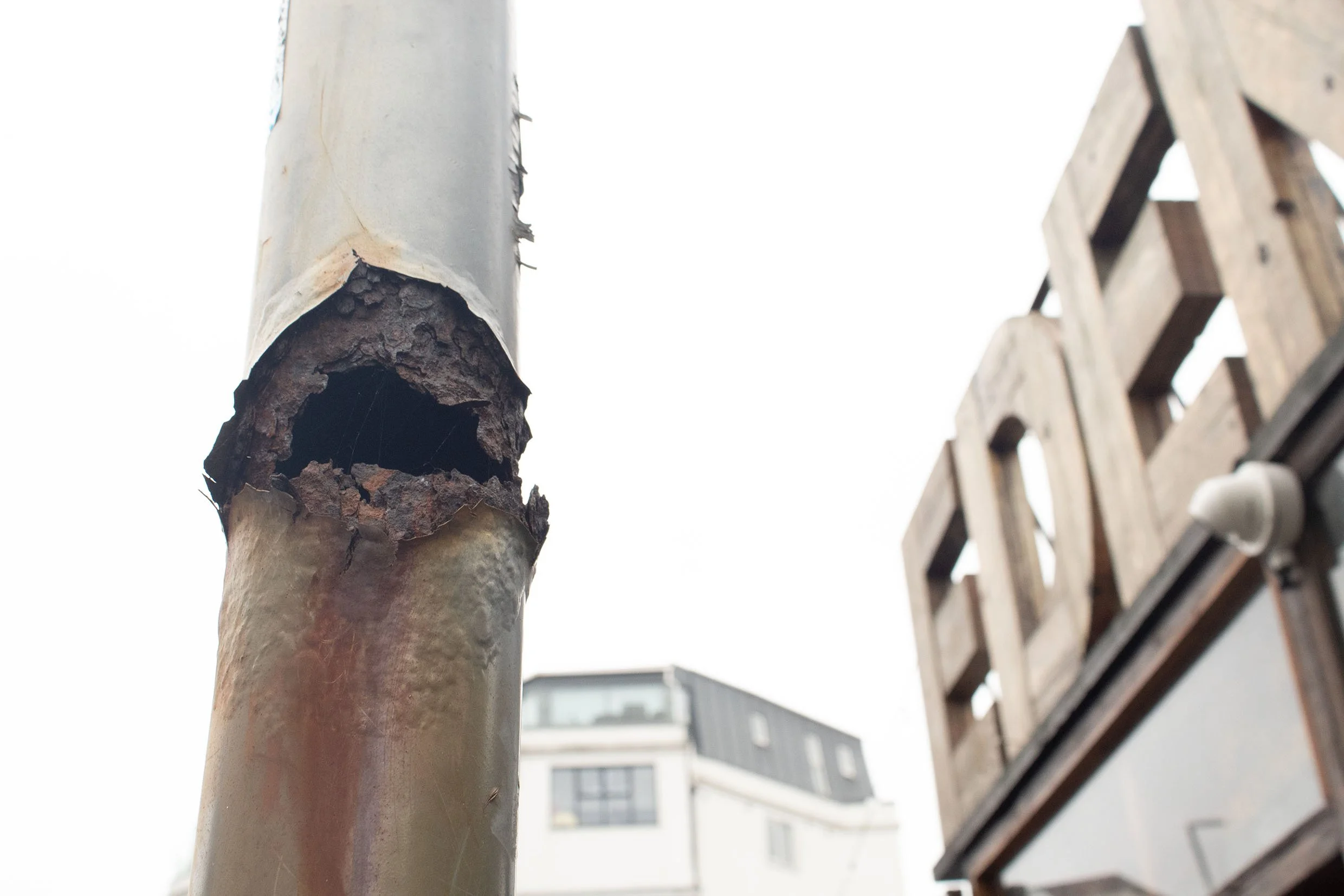 Close-up of a rusted, hollow metal pipe with a weathered surface and peeling paint, with a wooden lattice fence and apartment building in the background.
