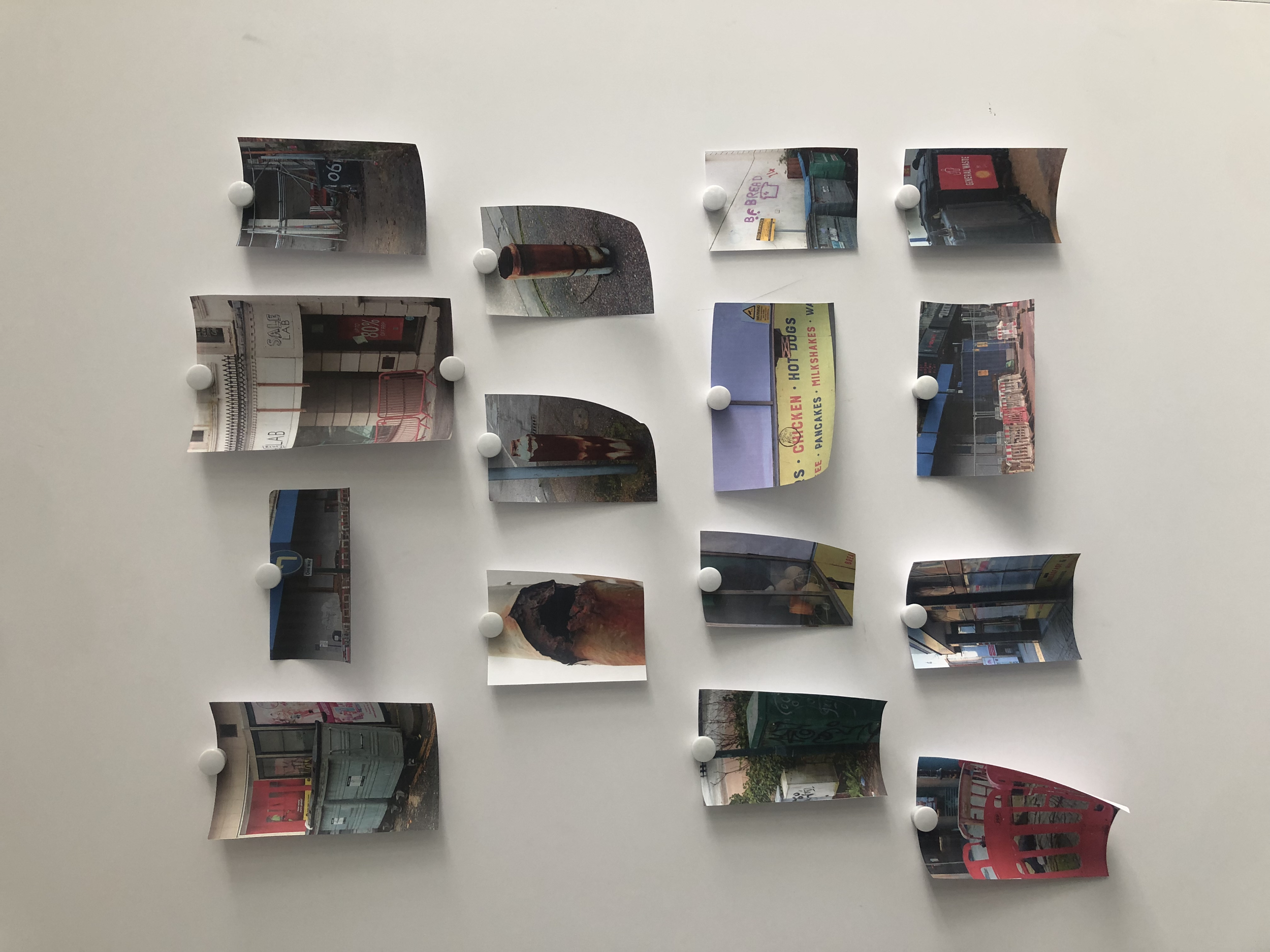 A white wall with several small printed photographs or images attached using white pushpins in a grid-like arrangement. The photos depict various scenes including city streets, storefronts, signs, and urban settings.
