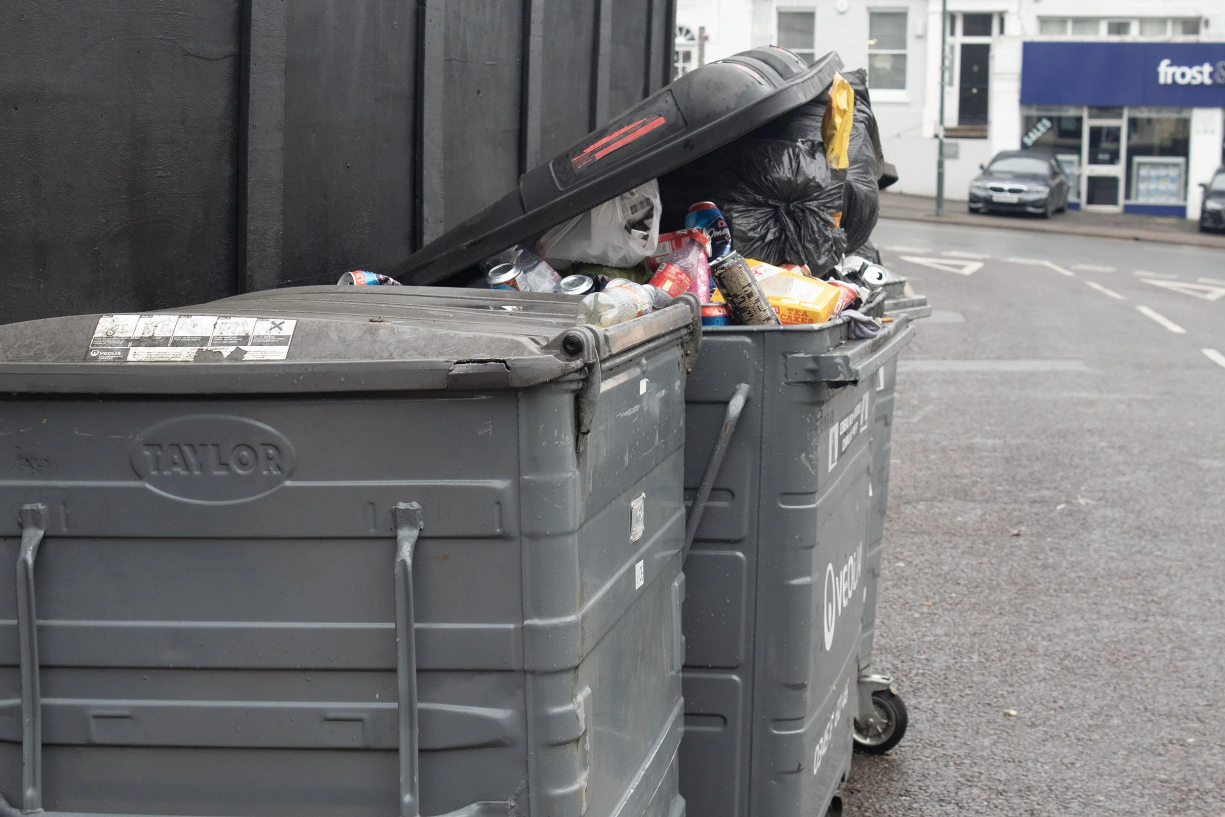 Two large gray trash bins filled with garbage on a city street, with one lid tilted and trash, including plastic bottles, cans, and black plastic bags overflowing. In the background are buildings and parked cars.