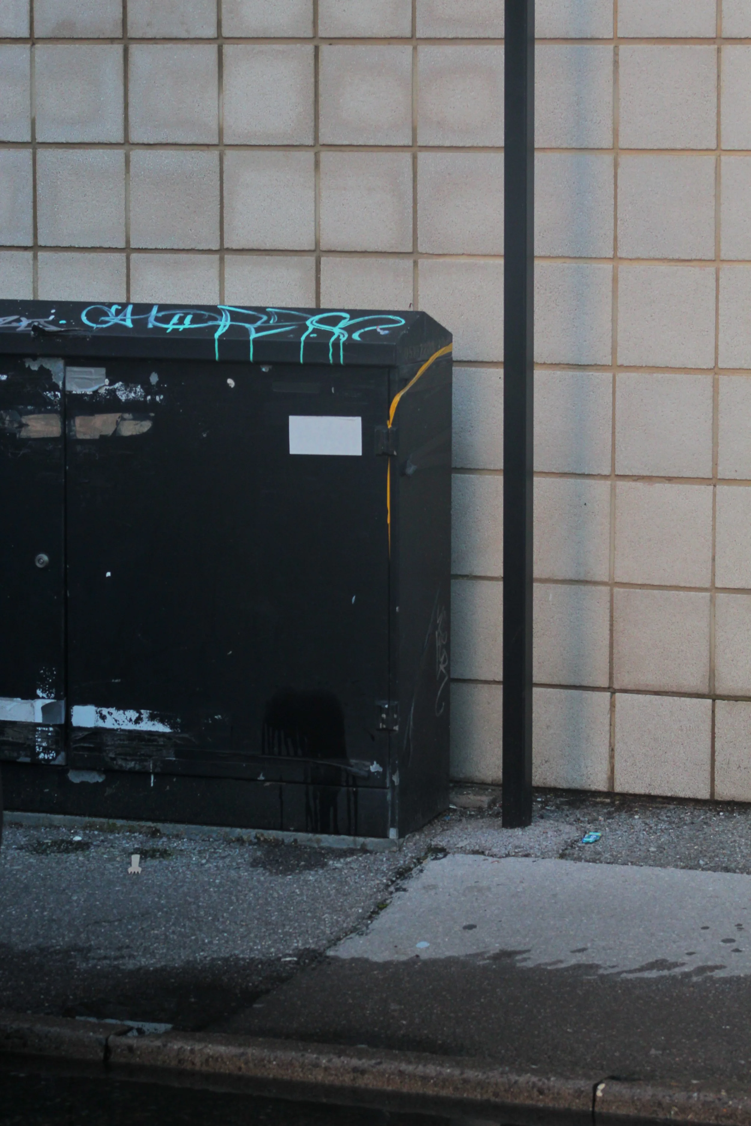 A black electrical box with graffiti on it, positioned next to a black metal pole, against a beige brick wall, on a rough asphalt ground.