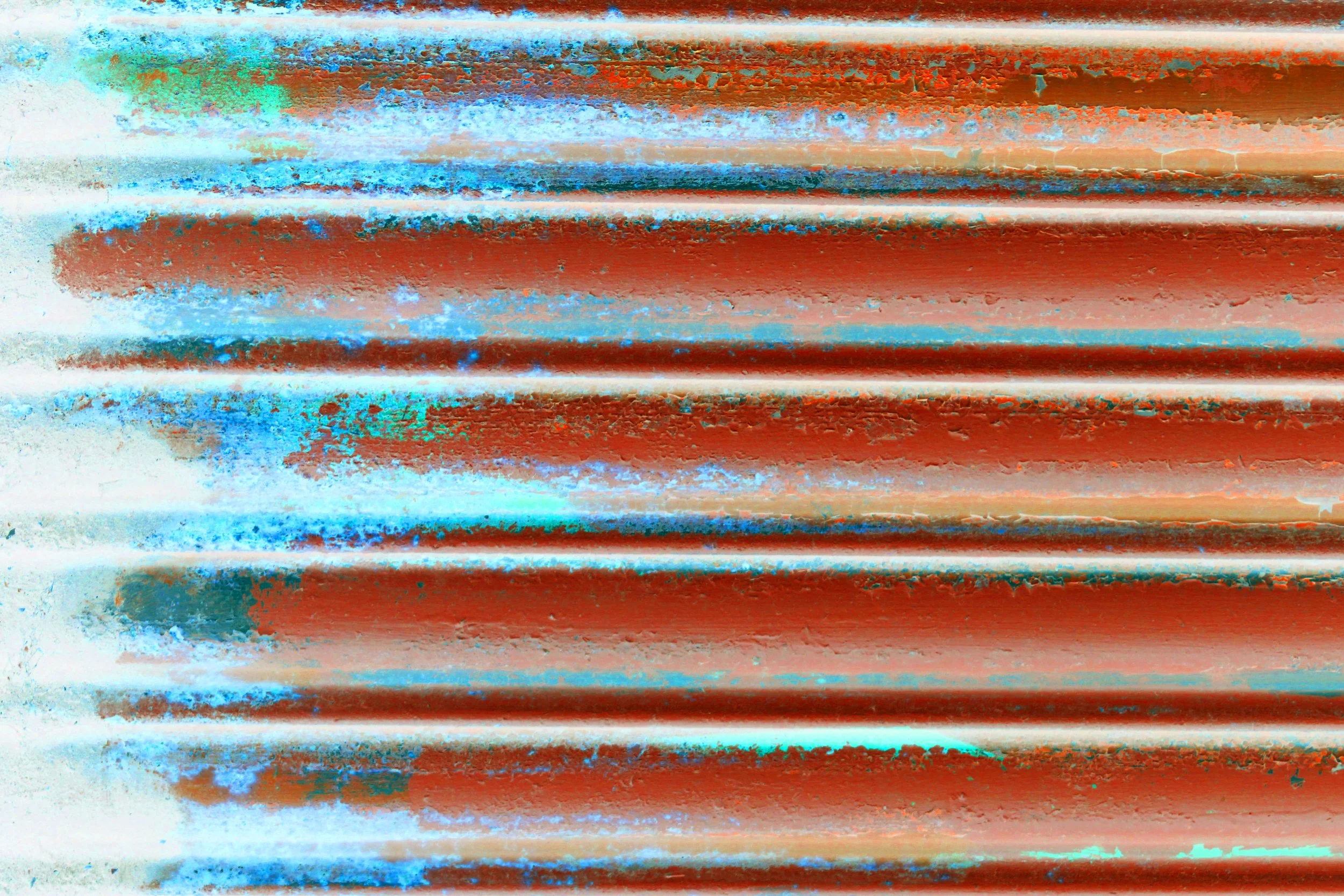 Close-up of a metallic surface with horizontal ridges, showing rust and peeling paint in shades of red, blue, and green, with textured and weathered appearance.