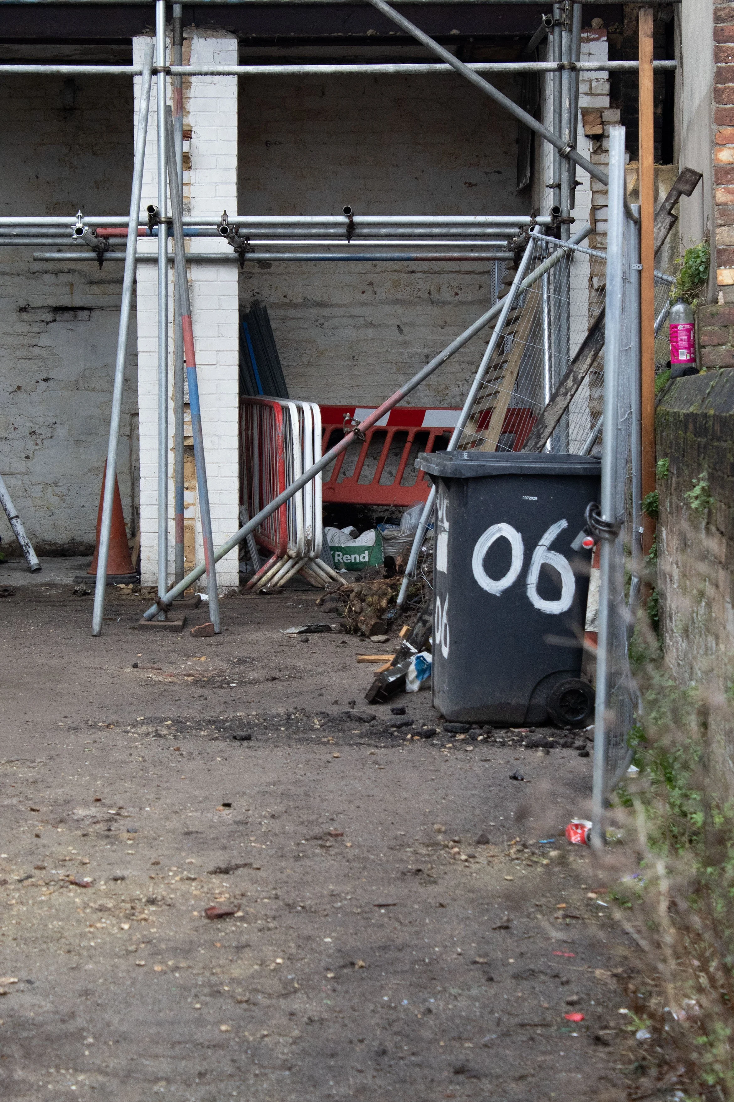 Construction site with scaffolding, orange and white safety barriers, a trash bin, and scattered debris on the ground.