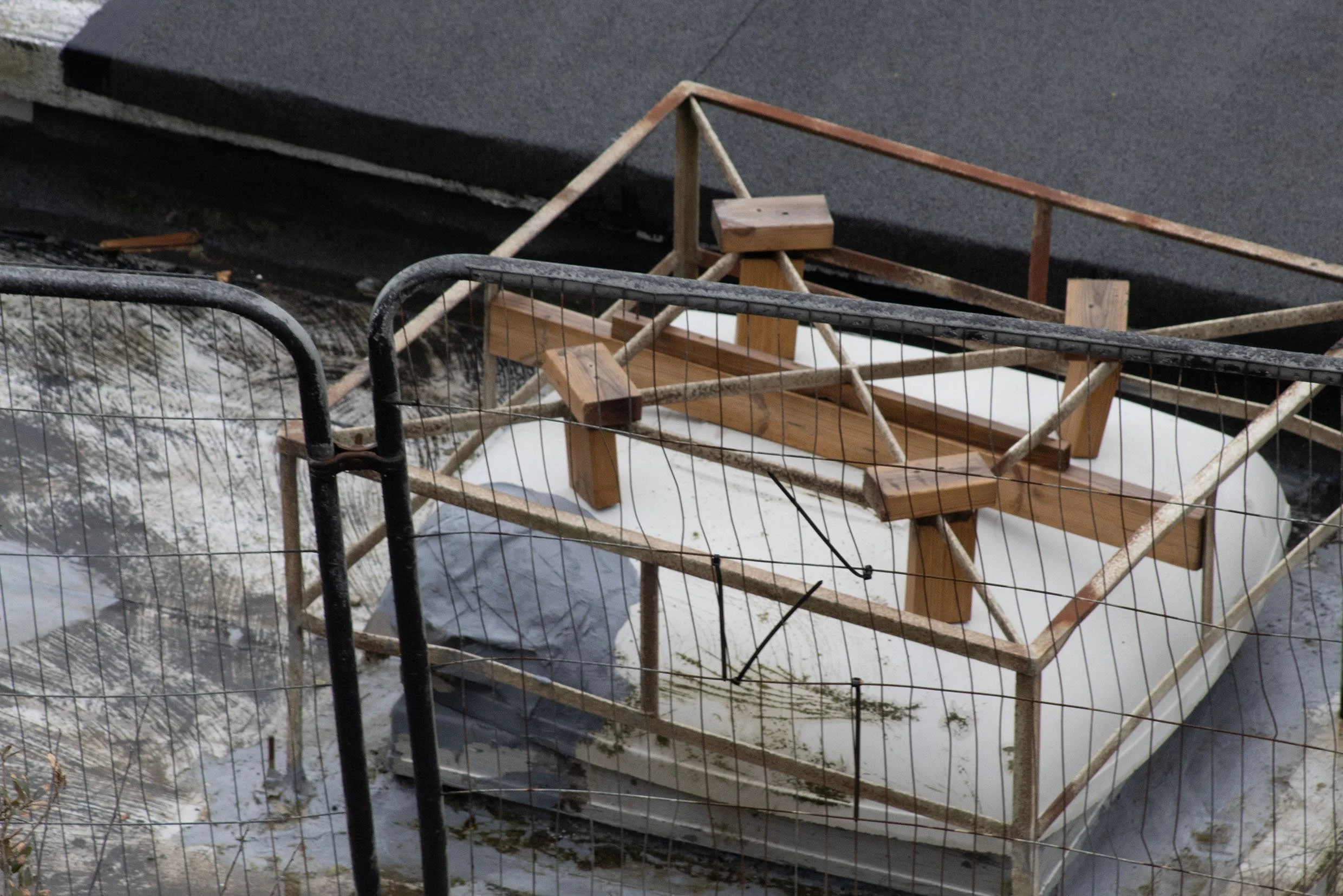A fallen wooden crib or playpen with a small wooden table inside, surrounded by metal fencing on a snowy ground with some patches of grass.