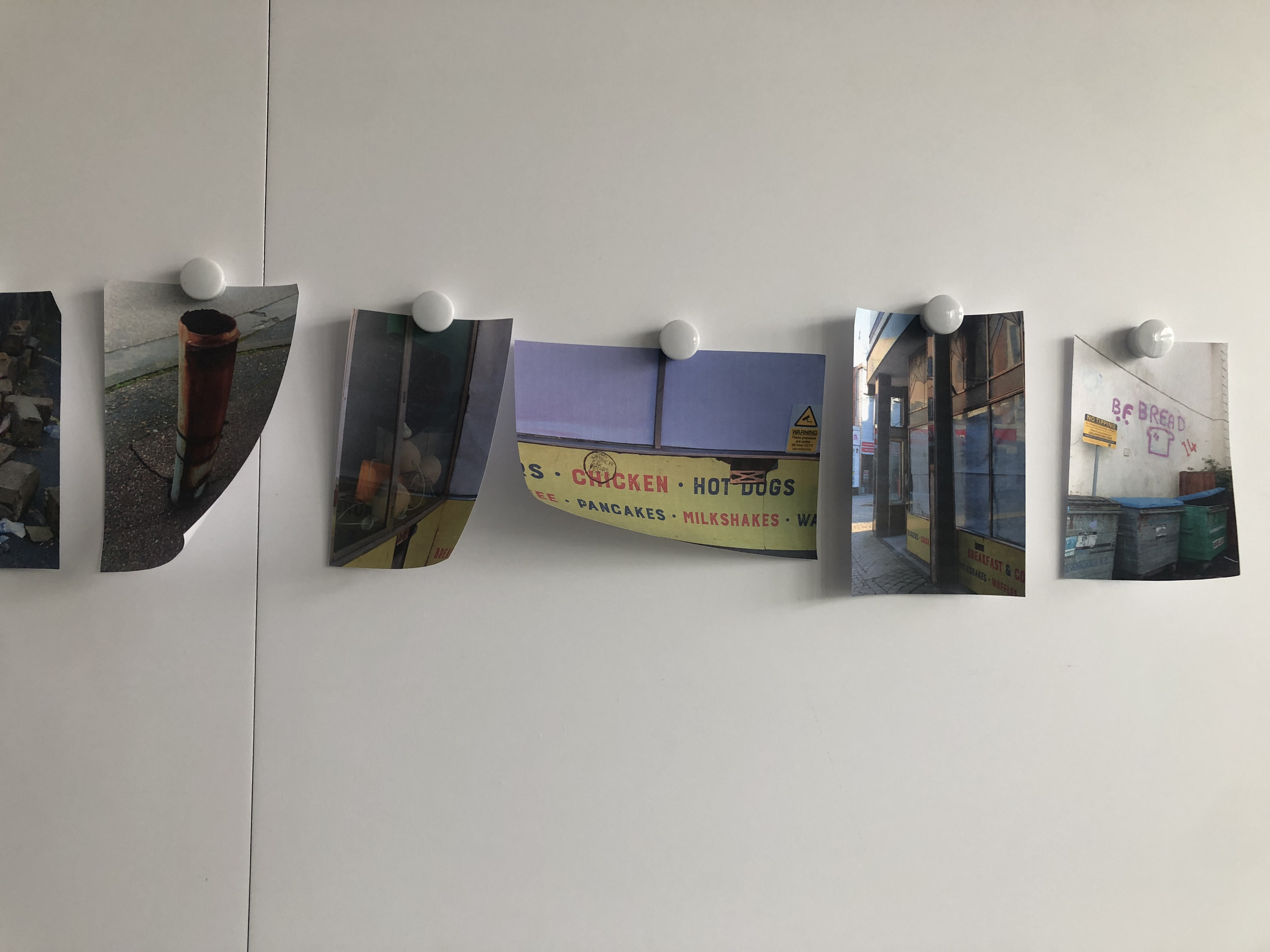 A row of photographs hanging on a white wall, held by white magnetic pins. The photographs include images of food, architecture, and signs.