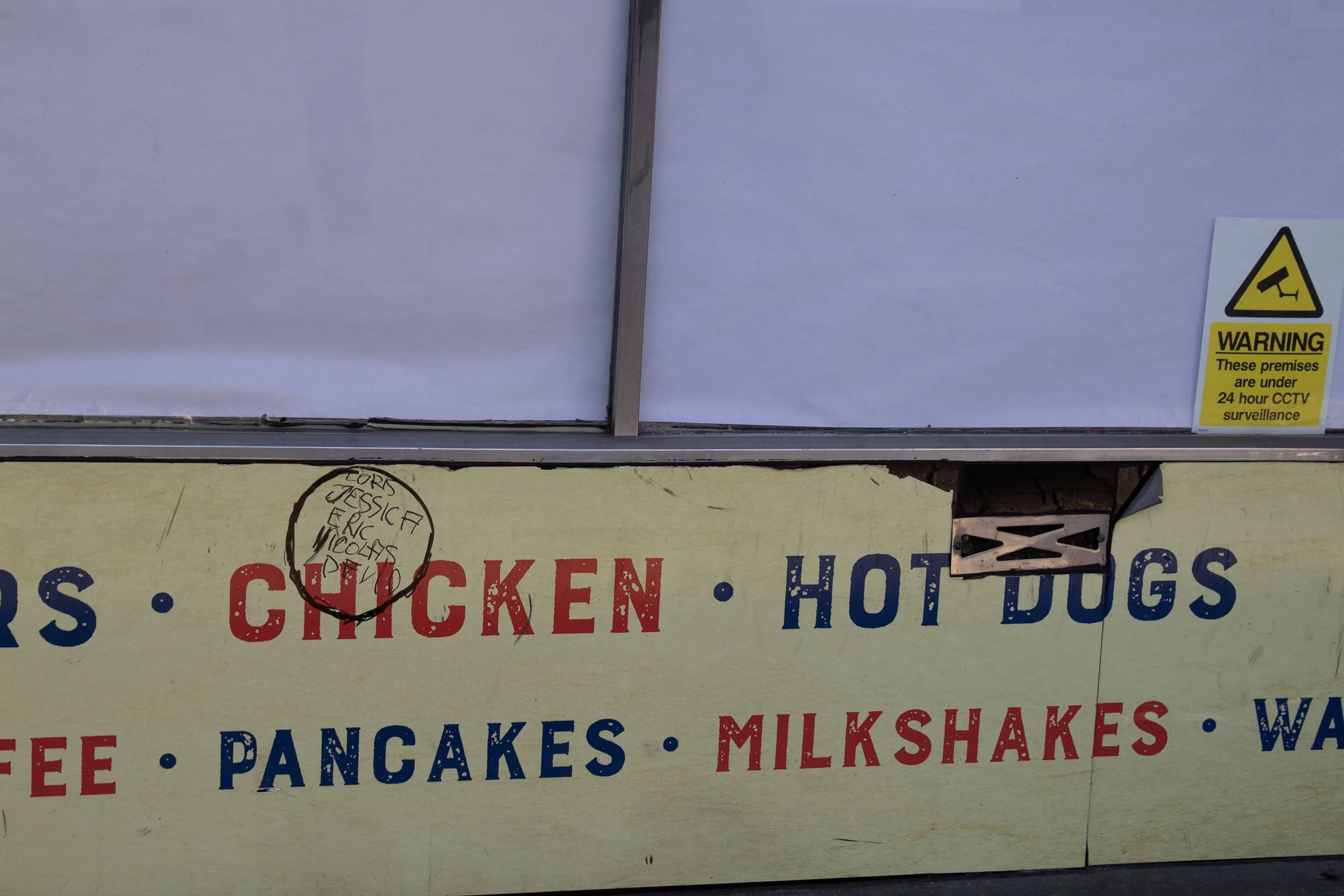 Part of a storefront sign advertising food items including chicken, hot dogs, pancakes, and milkshakes, with a warning sign about CCTV surveillance on the door.