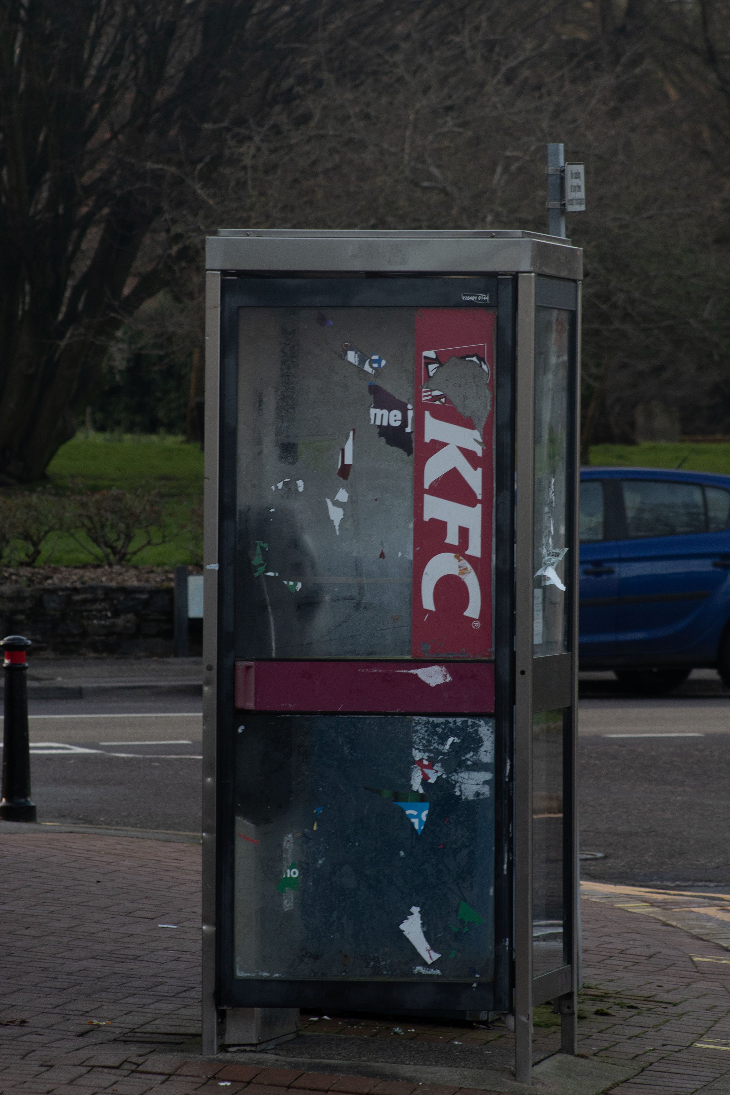 Empty, vandalized advertisement display case on a sidewalk with torn stickers and peeling posters, a street, parked cars, leafless trees, and a hedge in the background.