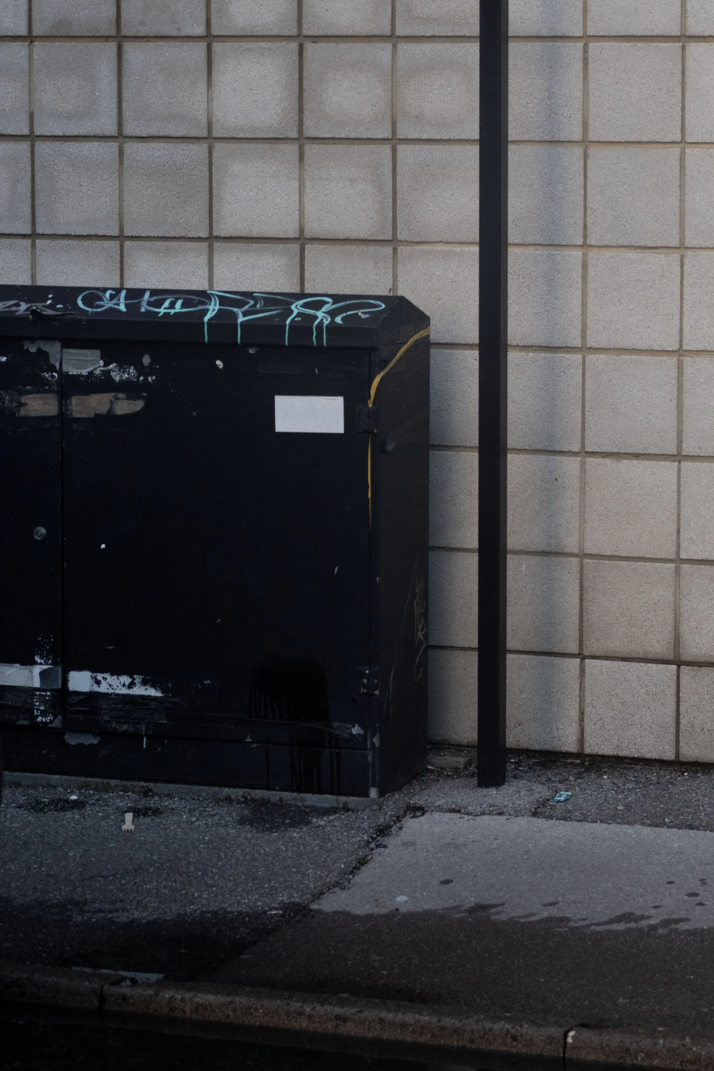 A black, graffiti-covered electrical box is situated against a beige tiled wall. There is a vertical black pole attached to the right side of the box. The ground is asphalt with some debris and a small puddle.