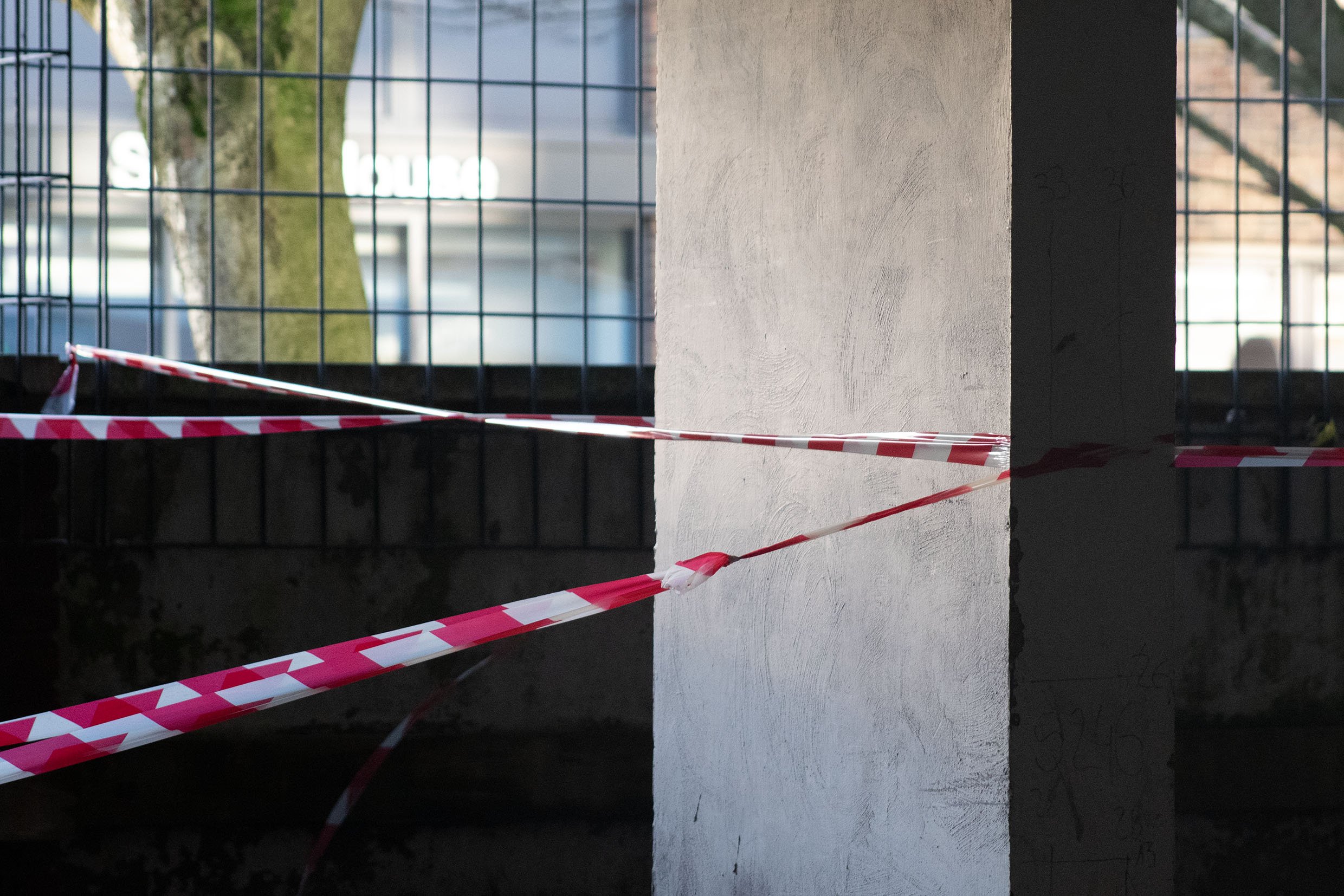 A construction site with a white concrete wall, a black fence, and red and white caution tape. In the background, there is a blurred building and a tree.