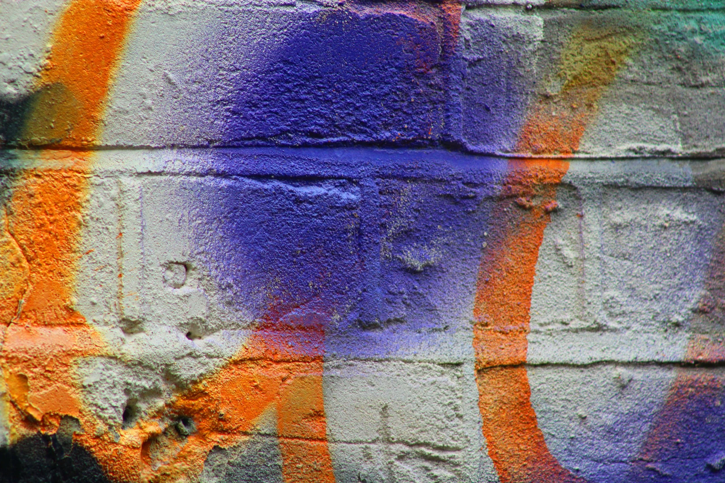 Close-up of a weathered brick wall with colorful spray paint splashes in purple, orange, and yellow.