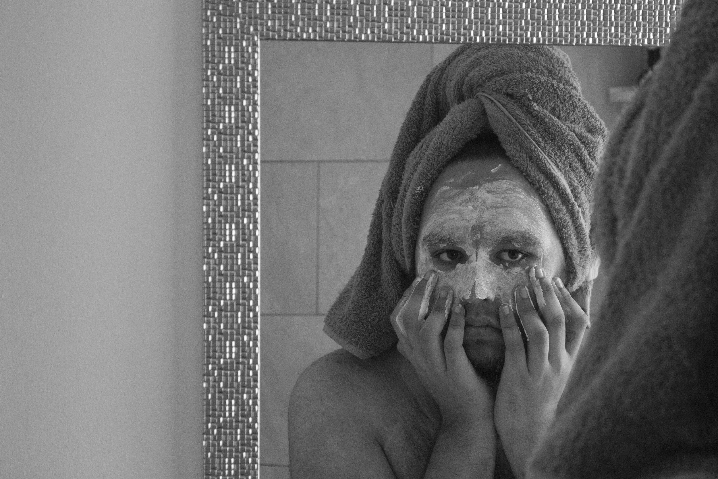 A woman with a towel wrapped around her head is applying a facial mask and looking into a mirror in a bathroom.