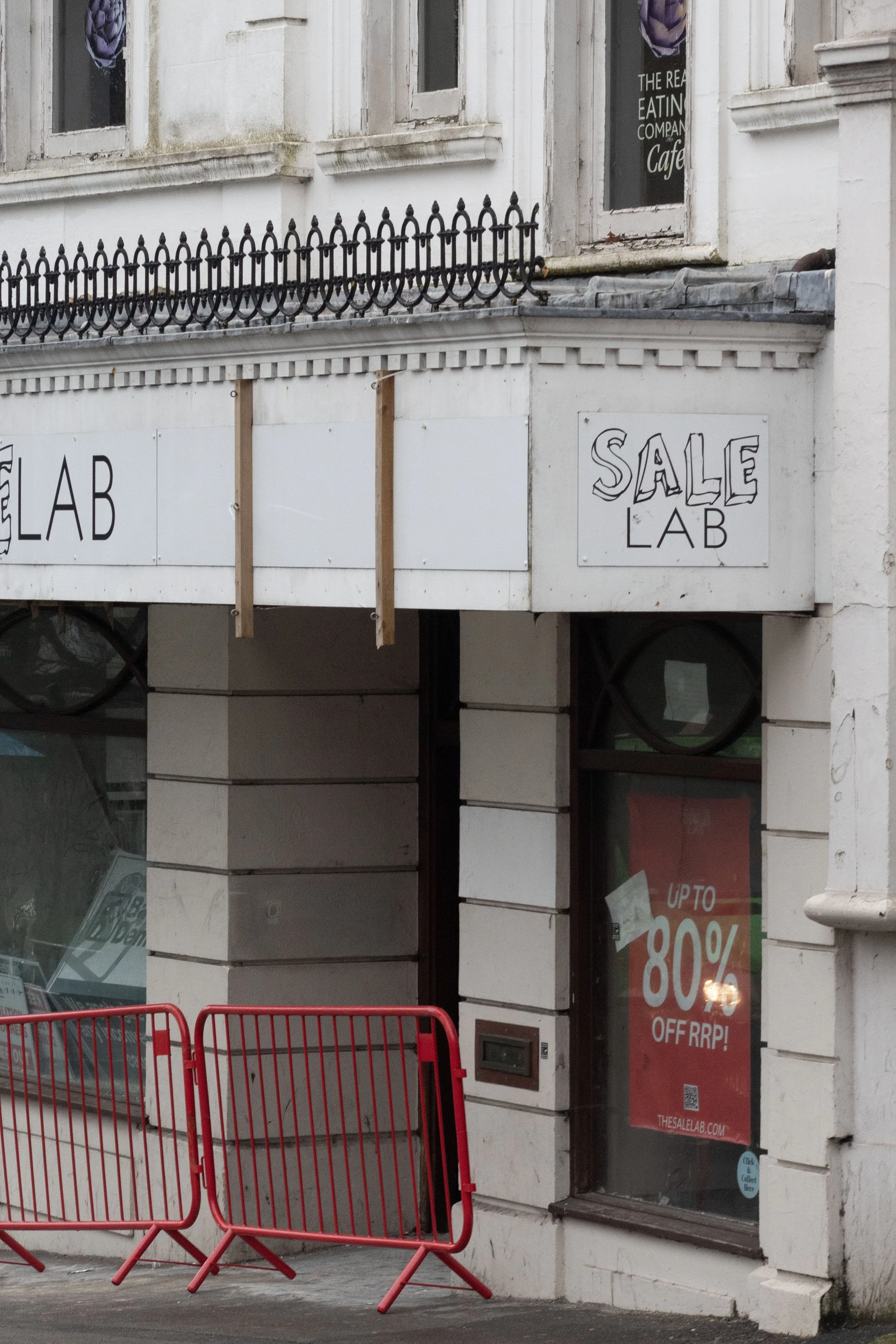 A storefront with signs for a sale and a cafe. Red barricades are placed outside, blocking part of the sidewalk. The building has white-painted brick walls and windows.