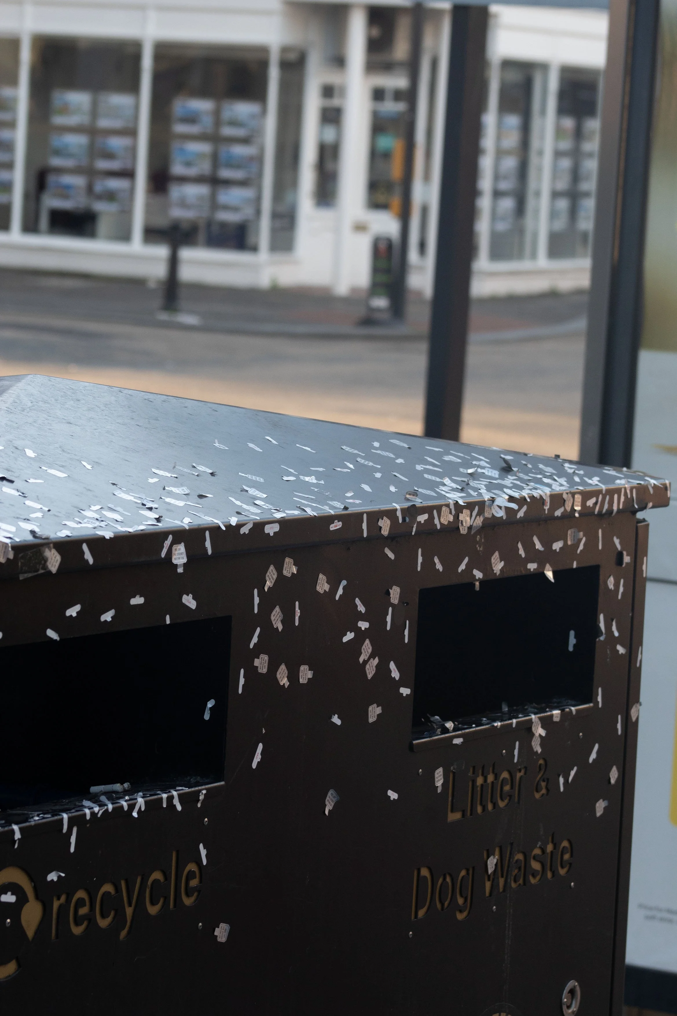 A black recycling bin with cut-out words 'recycle,' 'Dog waste,' and 'garbage' on it, decorated with numerous small white stickers, outdoors near a building with glass windows.