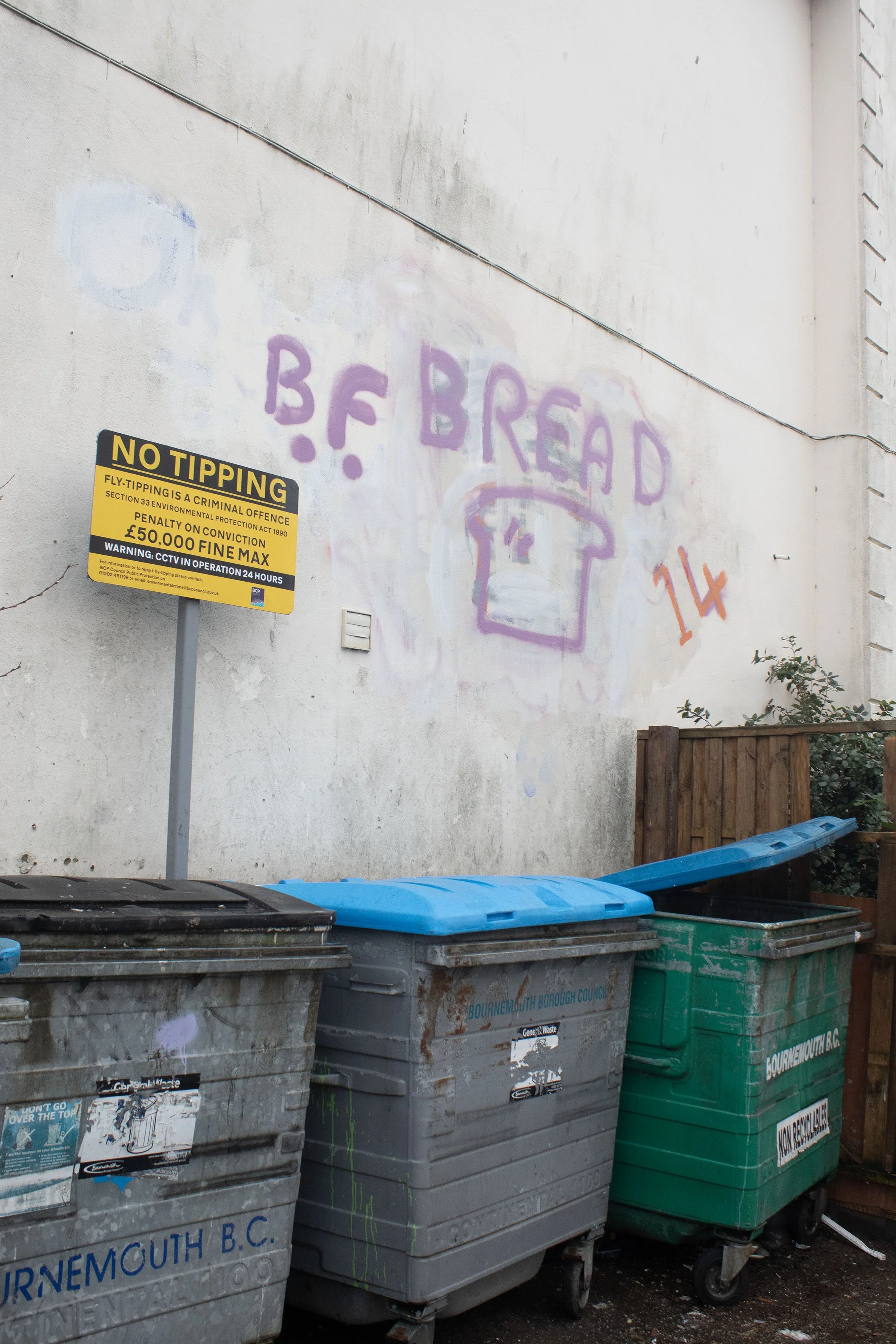 A white wall with graffiti text reading 'BREAD' and a purple smiley face. There are four garbage bins at the bottom, two gray and two green, with black and blue lids. A yellow warning sign about no tipping is attached to a pole near the bins, and the