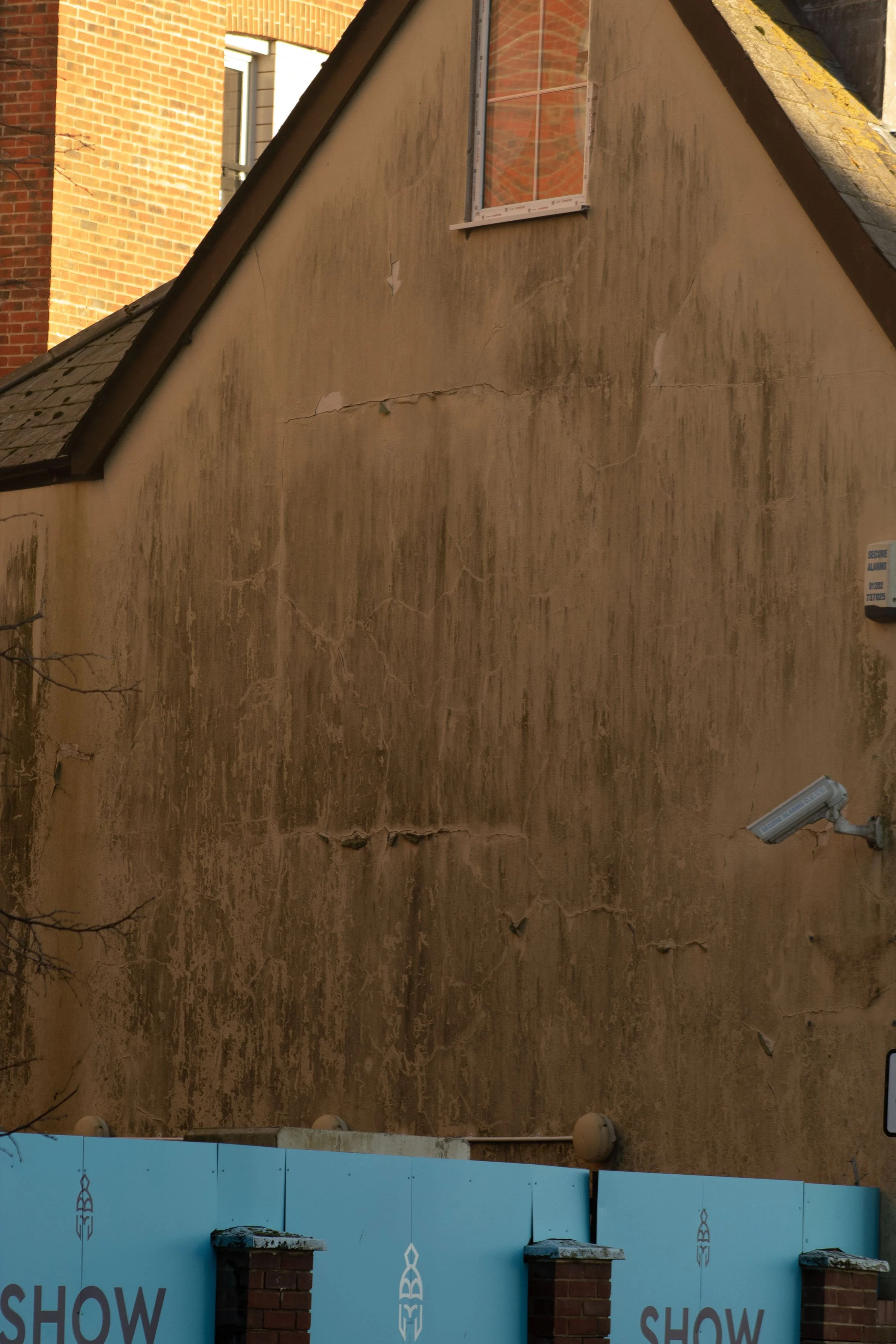Close-up view of a weathered beige wall with cracks and peeling paint, including a window at the top with reflective glass showing a brick building.