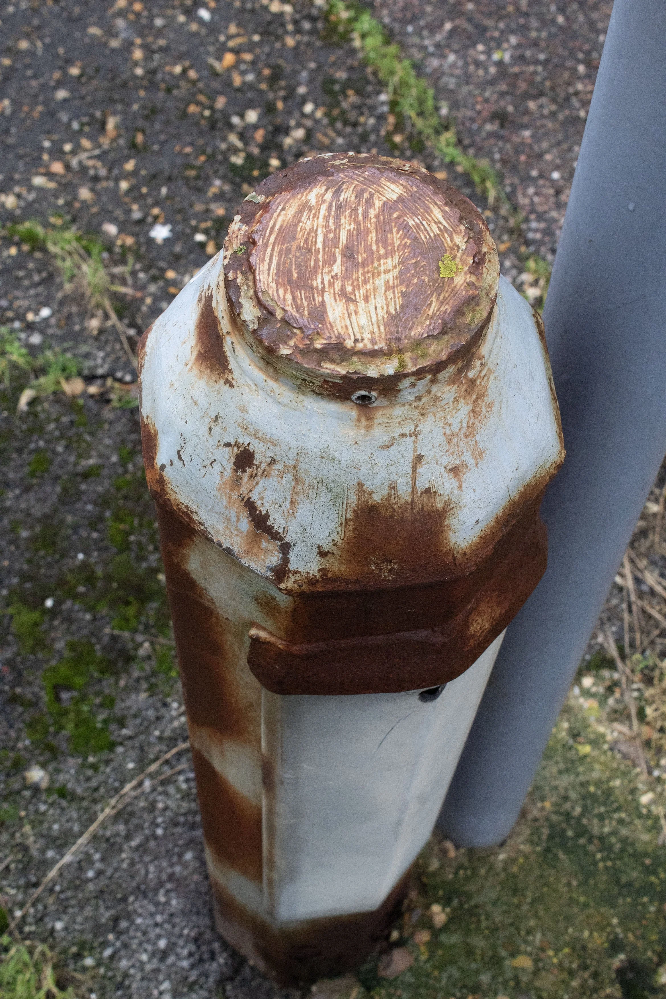 Close-up of a parking barrier post with rust and peeling paint, situated outdoors on an asphalt surface with some patches of grass.