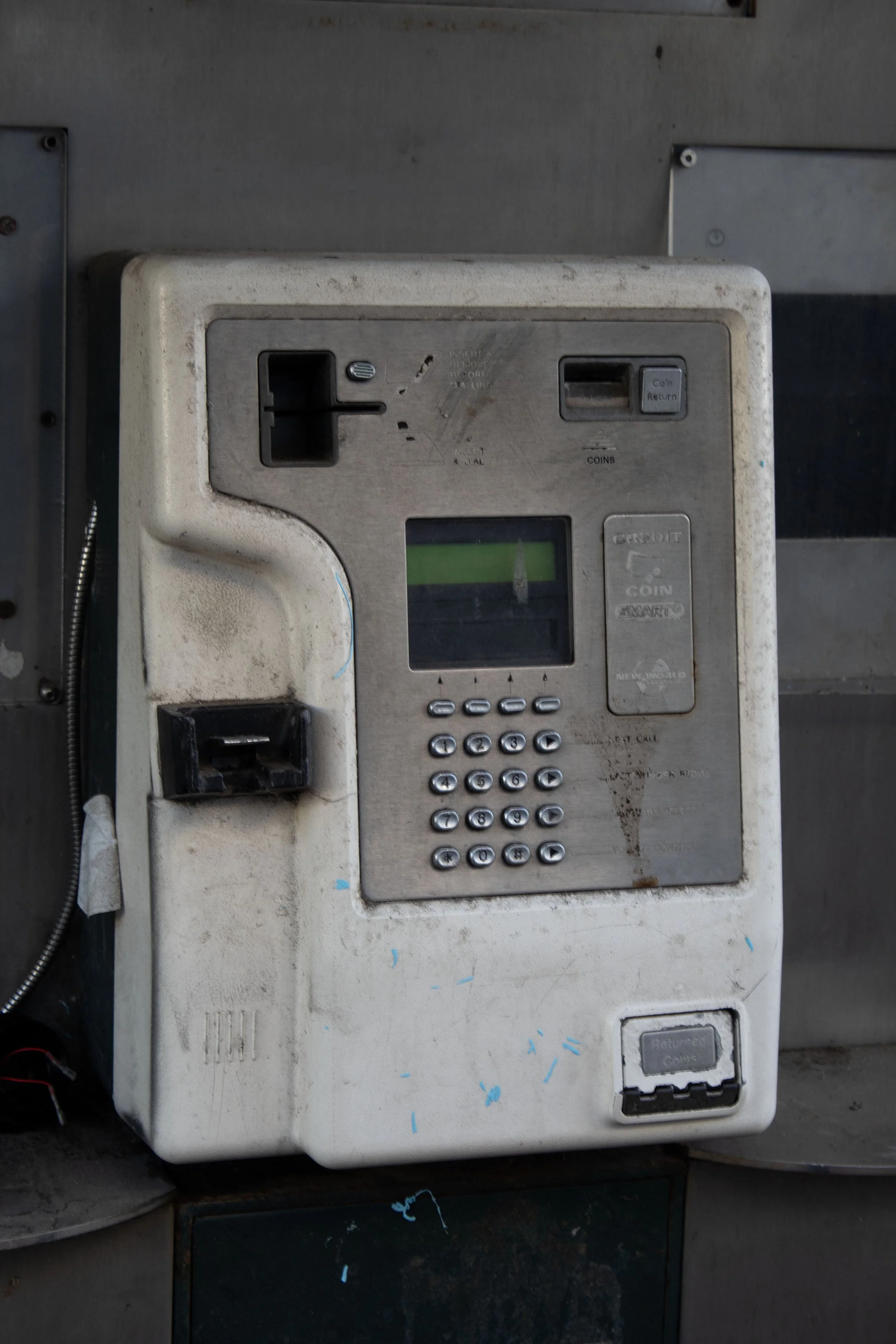 An old, dirty payphone mounted on a wall with a keypad, coin slot, and a card reader. The payphone appears worn out with visible dirt and scratches.
