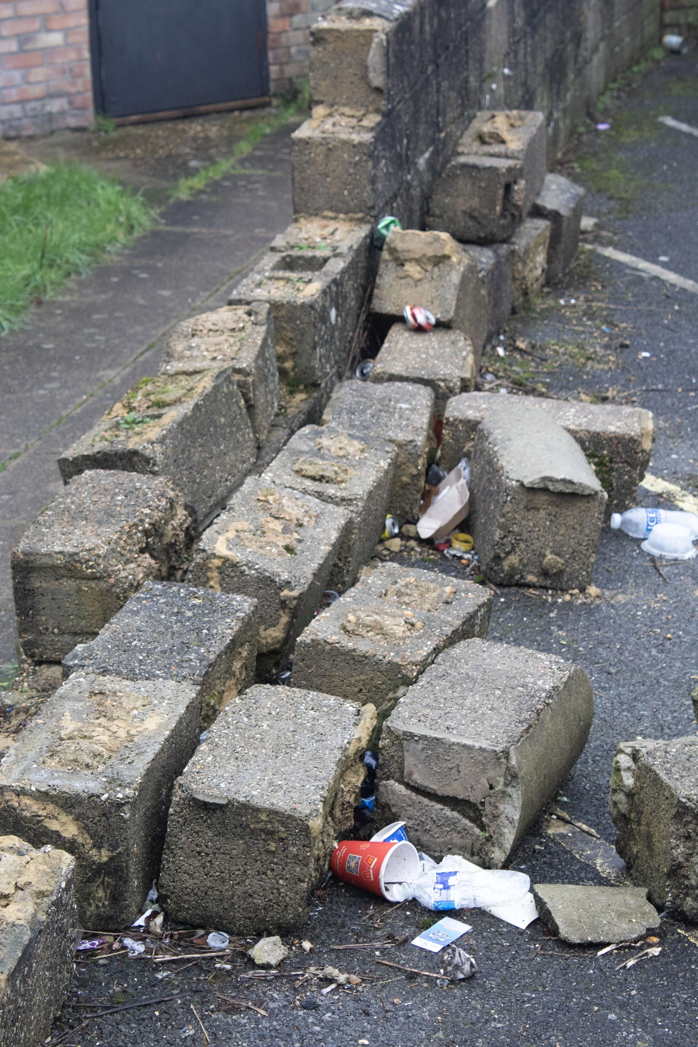 A section of sidewalk and a wall with concrete blocks placed along the edge, with trash such as paper cups, bottles, and miscellaneous debris scattered on the ground.