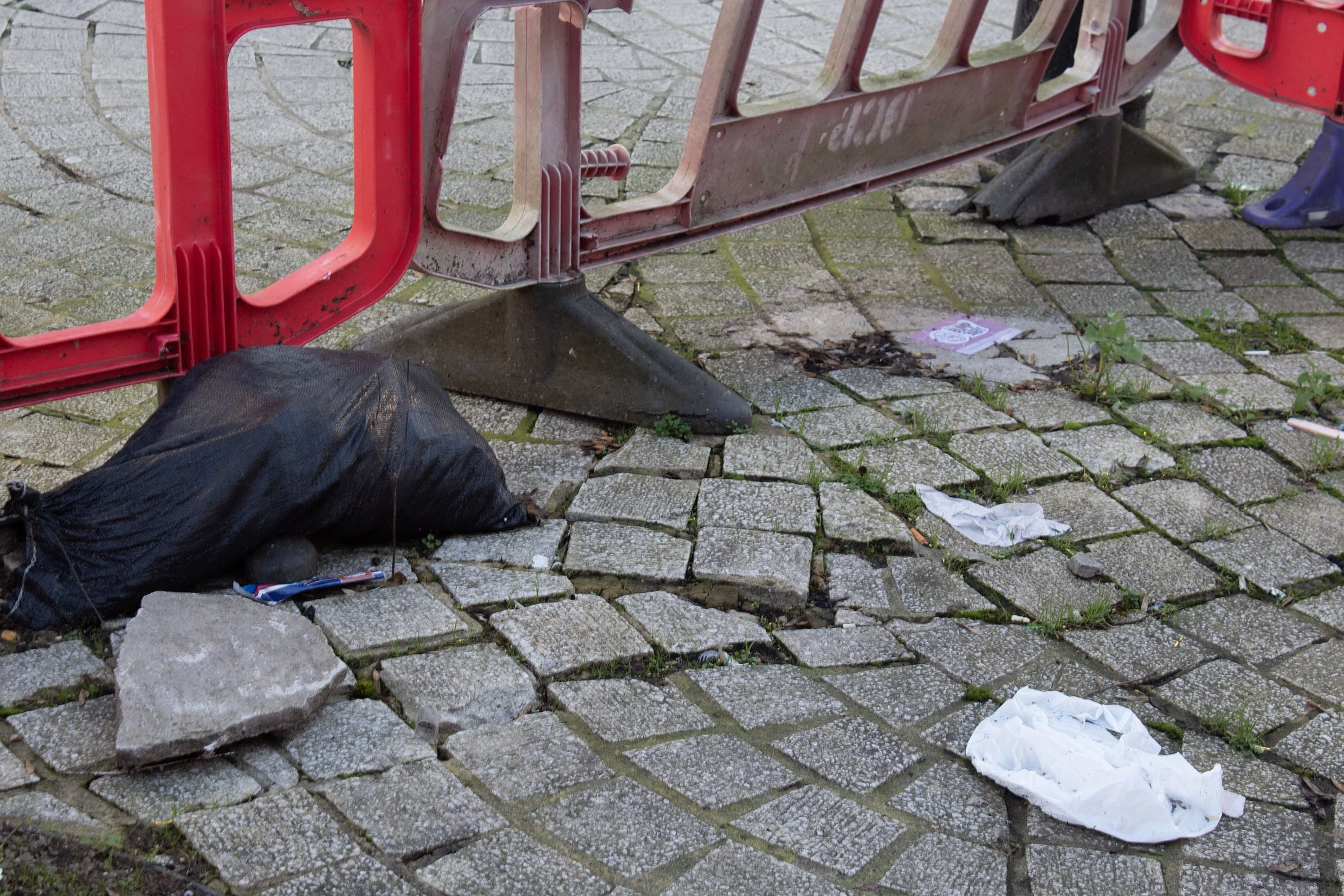 A sidewalk with broken and uneven cobblestones, a red plastic barrier, and scattered trash including a black bag, a crumpled paper, and a white tissue.