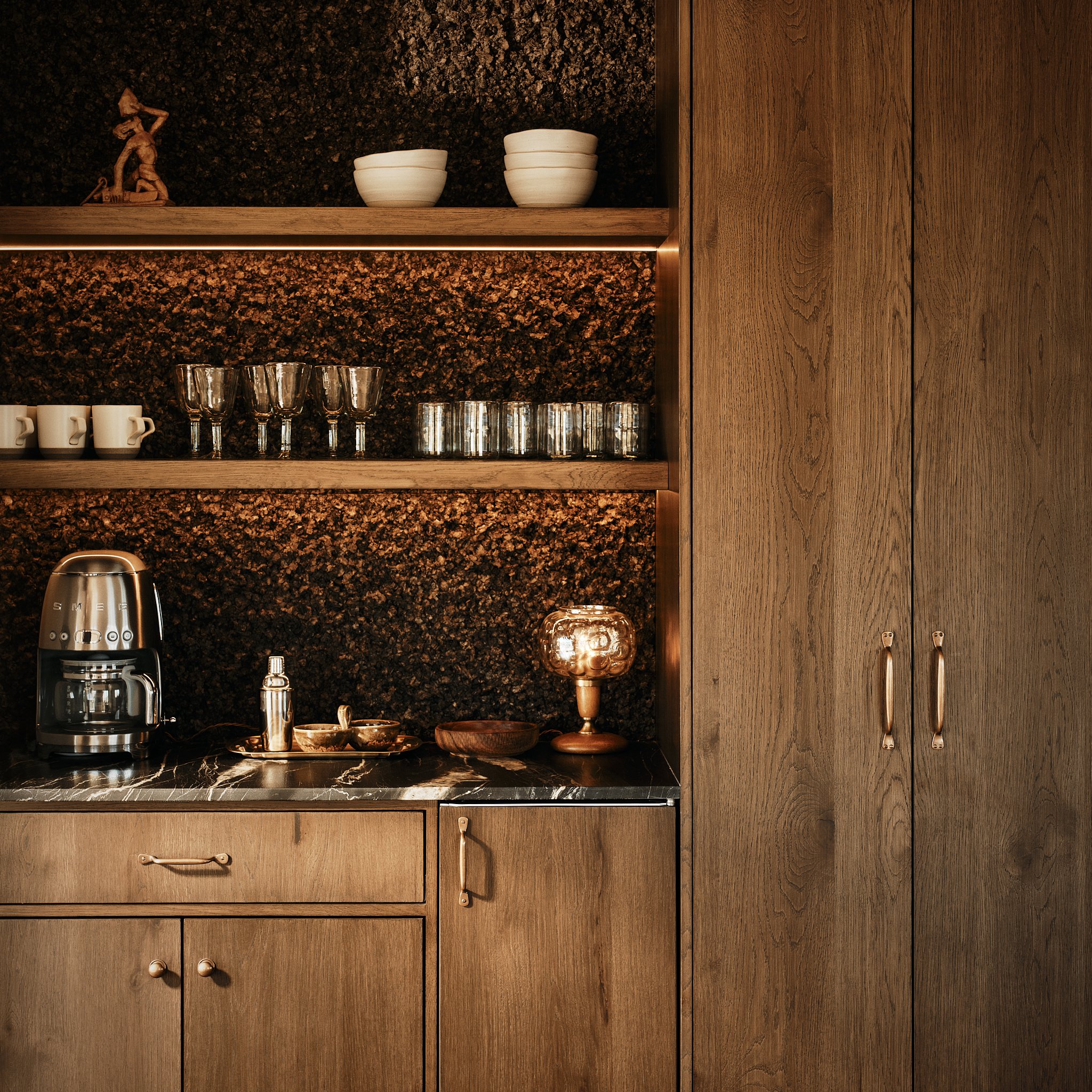 A kitchen with wooden cabinets, a shelf with white bowls, a small sculpture, and glassware including cups, wine glasses, and tumblers on black countertops.