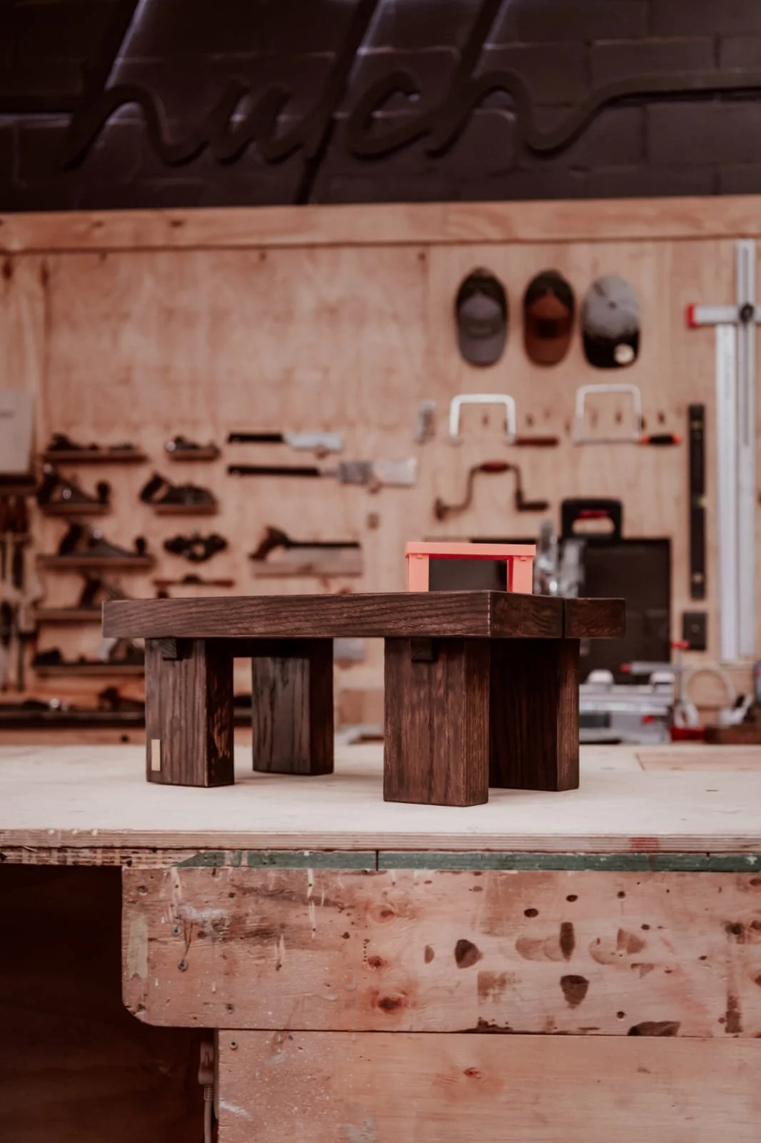 A small handcrafted wooden table in a woodworking shop, with a workbench and tool wall with various tools and hats in the background.