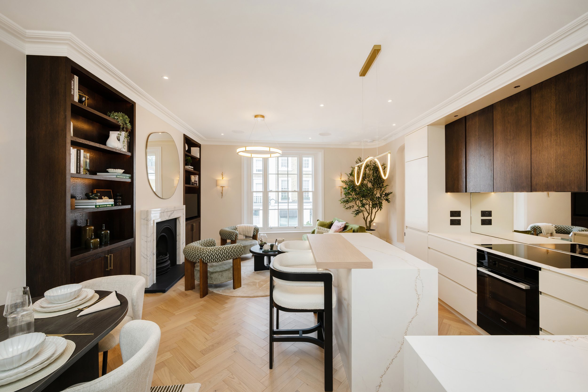Open-concept living room and kitchen with white walls, wooden floors, dark wood shelving, a fireplace, large window, modern lighting fixtures, and a green plant.