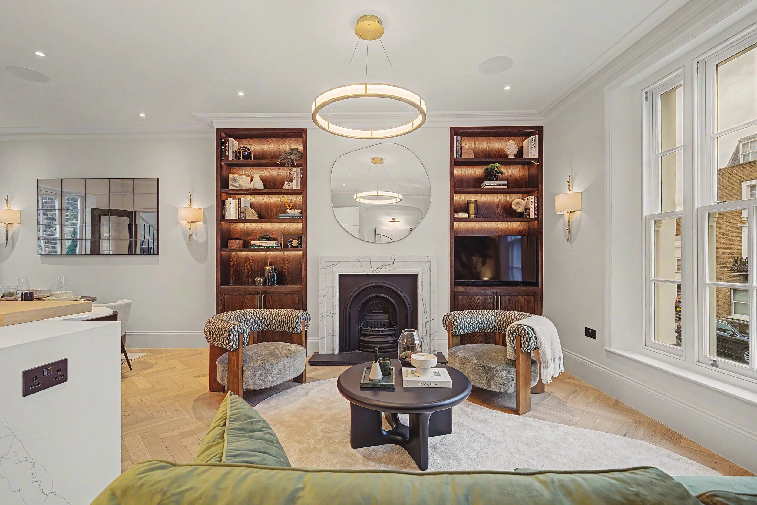 Living room with fireplace, wooden bookshelves, a circular black coffee table, striped upholstered chairs, large windows, and modern chandelier lighting.