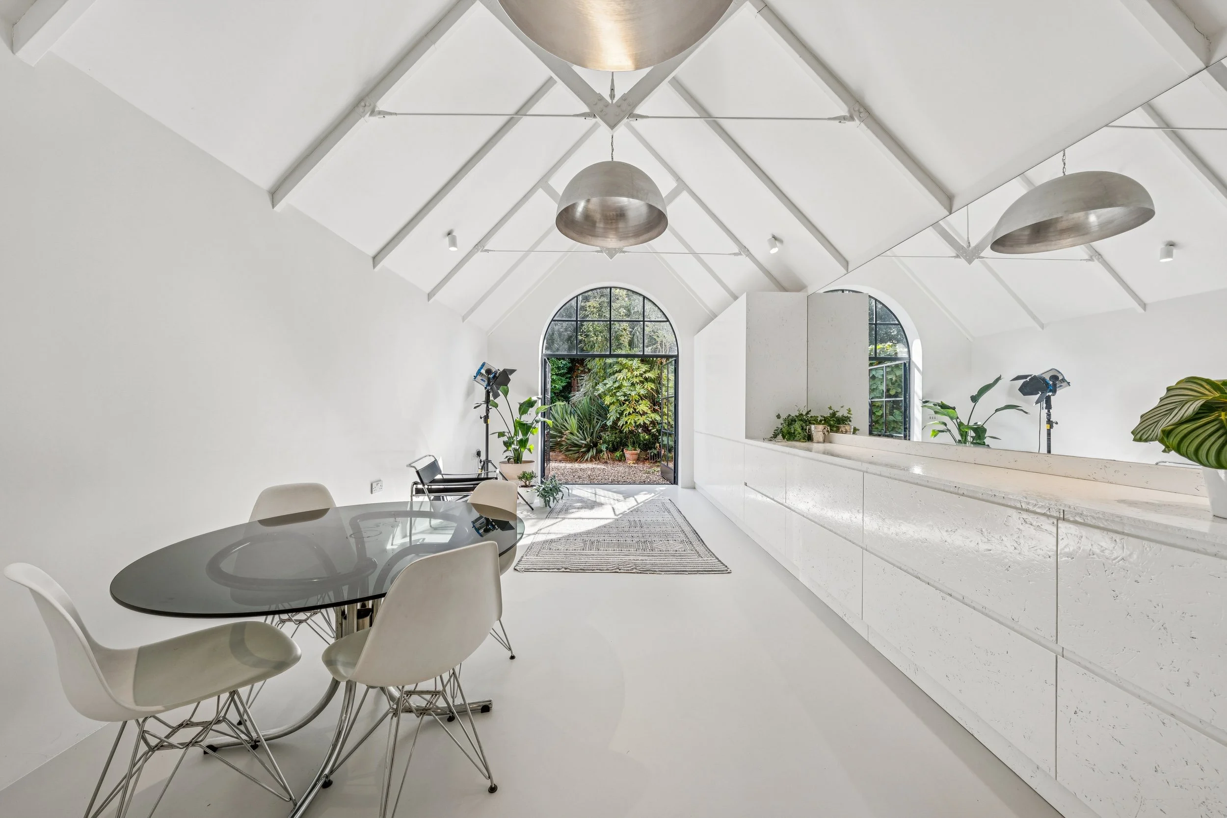 Modern white interior space with large arched windows, hanging light fixtures, a round dining table with chairs, and lush greenery outside.