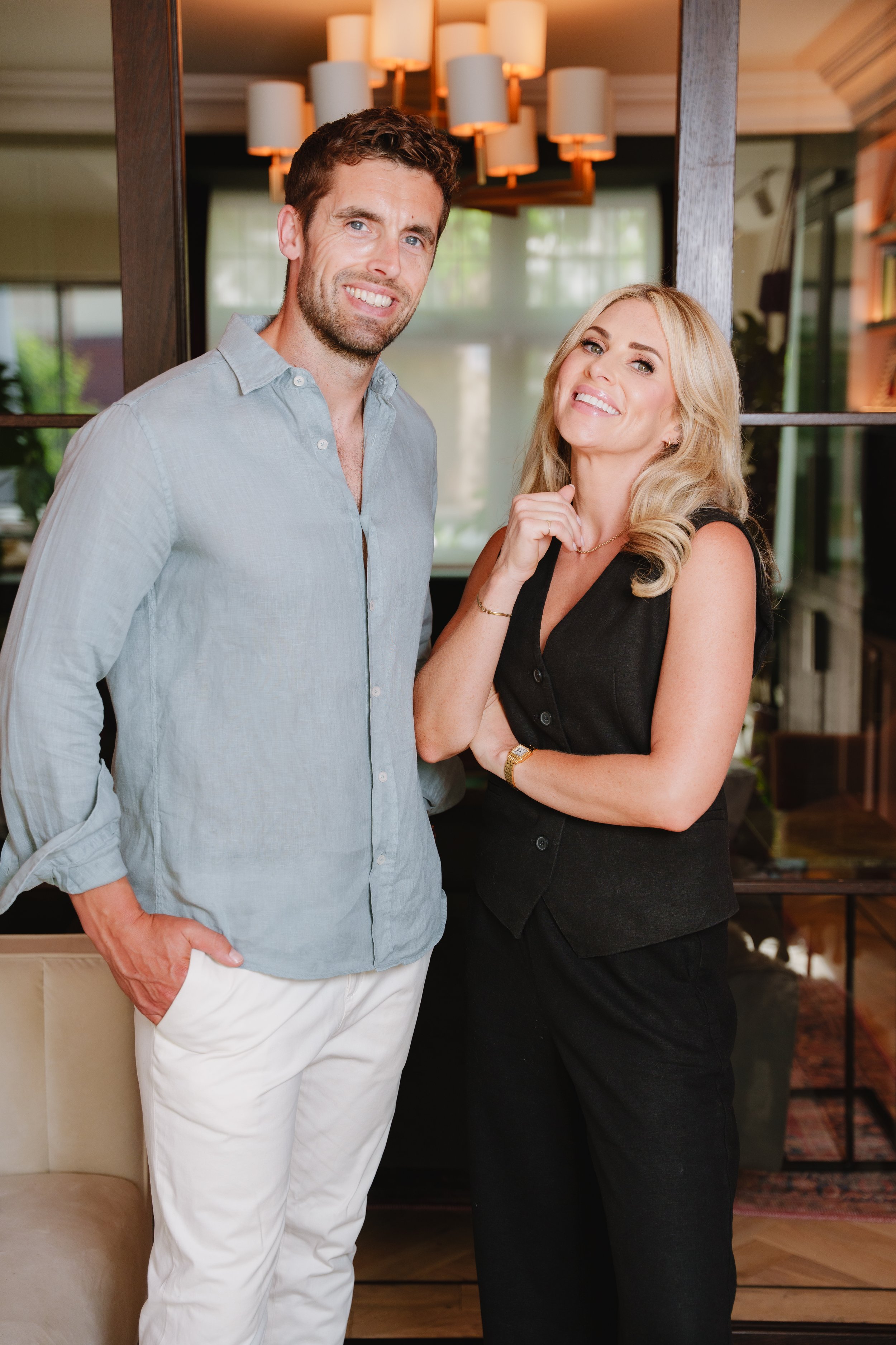 A man and woman smile and pose indoors near a wooden partition with glass panes and a bookshelf in the background. The man wears a light gray button-up shirt and white pants, the woman wears a black sleeveless top and black pants, both have light-colored hair.