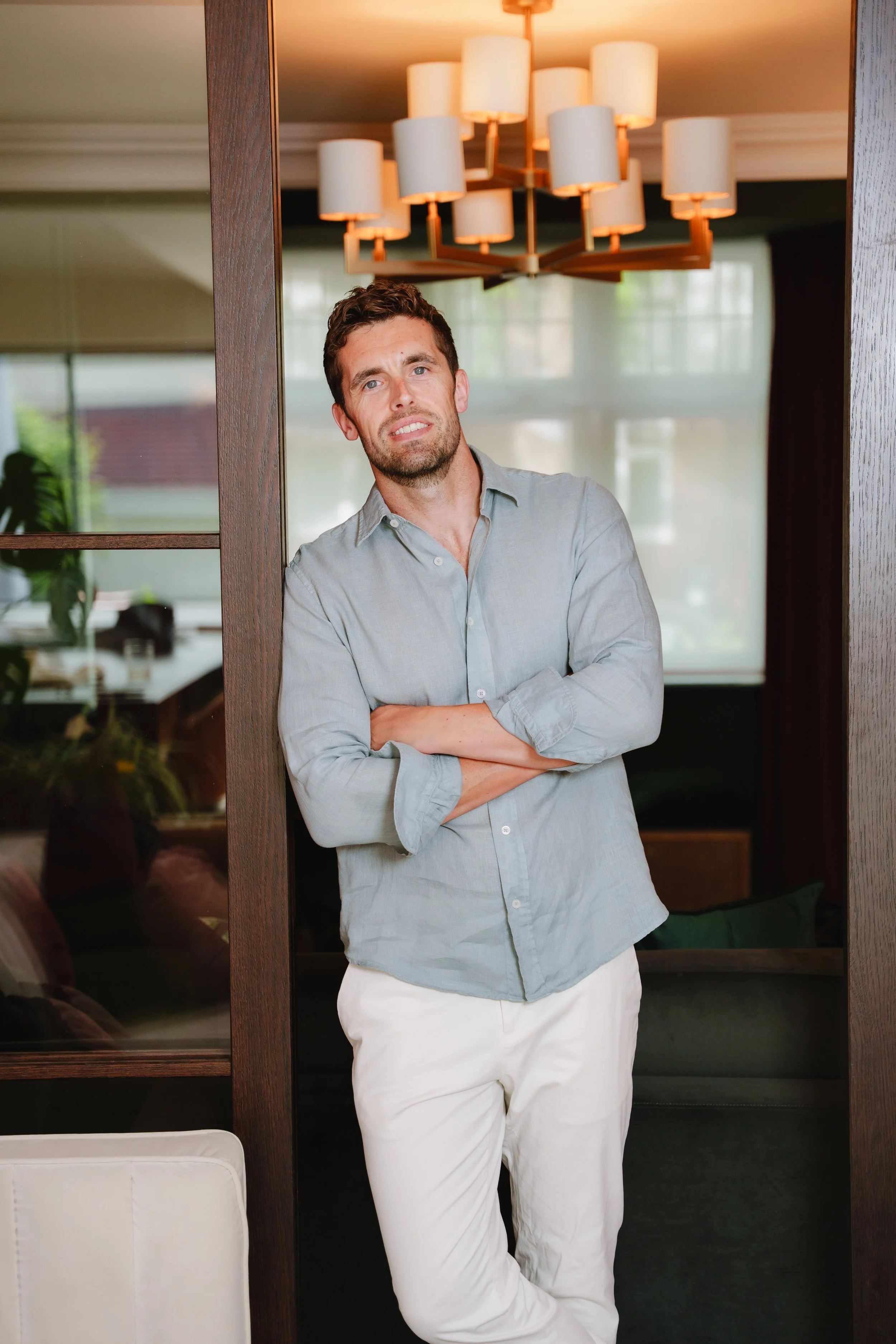 A man with brown hair and a beard, wearing a light gray button-up shirt and white pants, leaning against a wooden frame with his arms crossed, standing inside a room with large windows and a modern chandelier overhead.