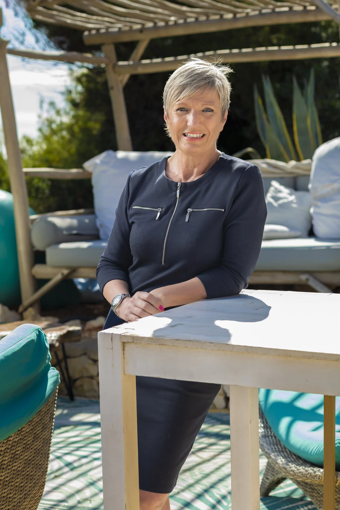 A middle-aged woman with short fiar hair wearing a blue zip-up dress, smiling while leaning on a table outdoors on a sunny day. There are outdoor furniture and greenery in the background.