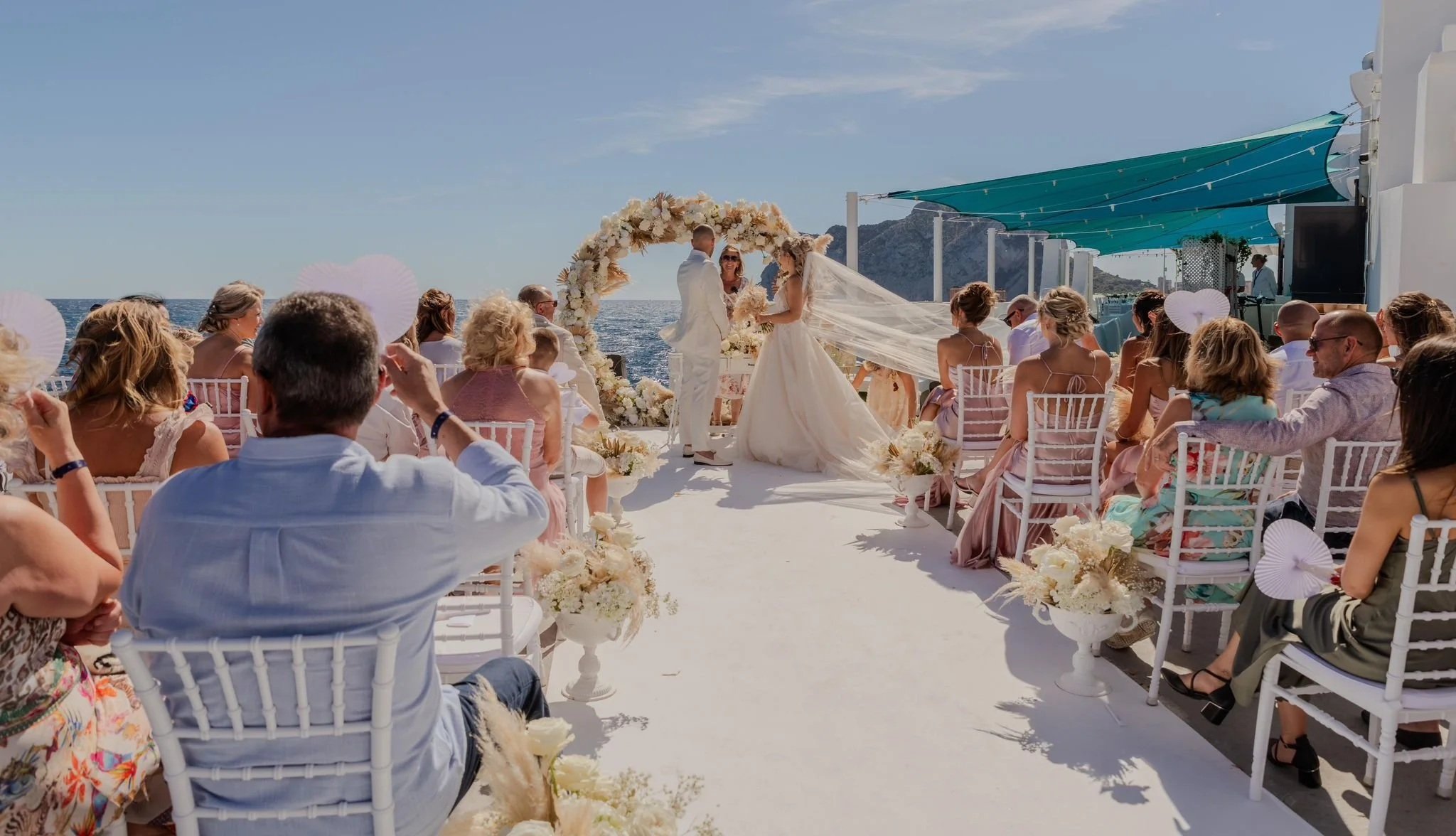 A wedding ceremony taking place on a boat with the ocean and mountains in the background. The bride and groom are exchanging vows under a floral arch, with guests seated on white chairs.