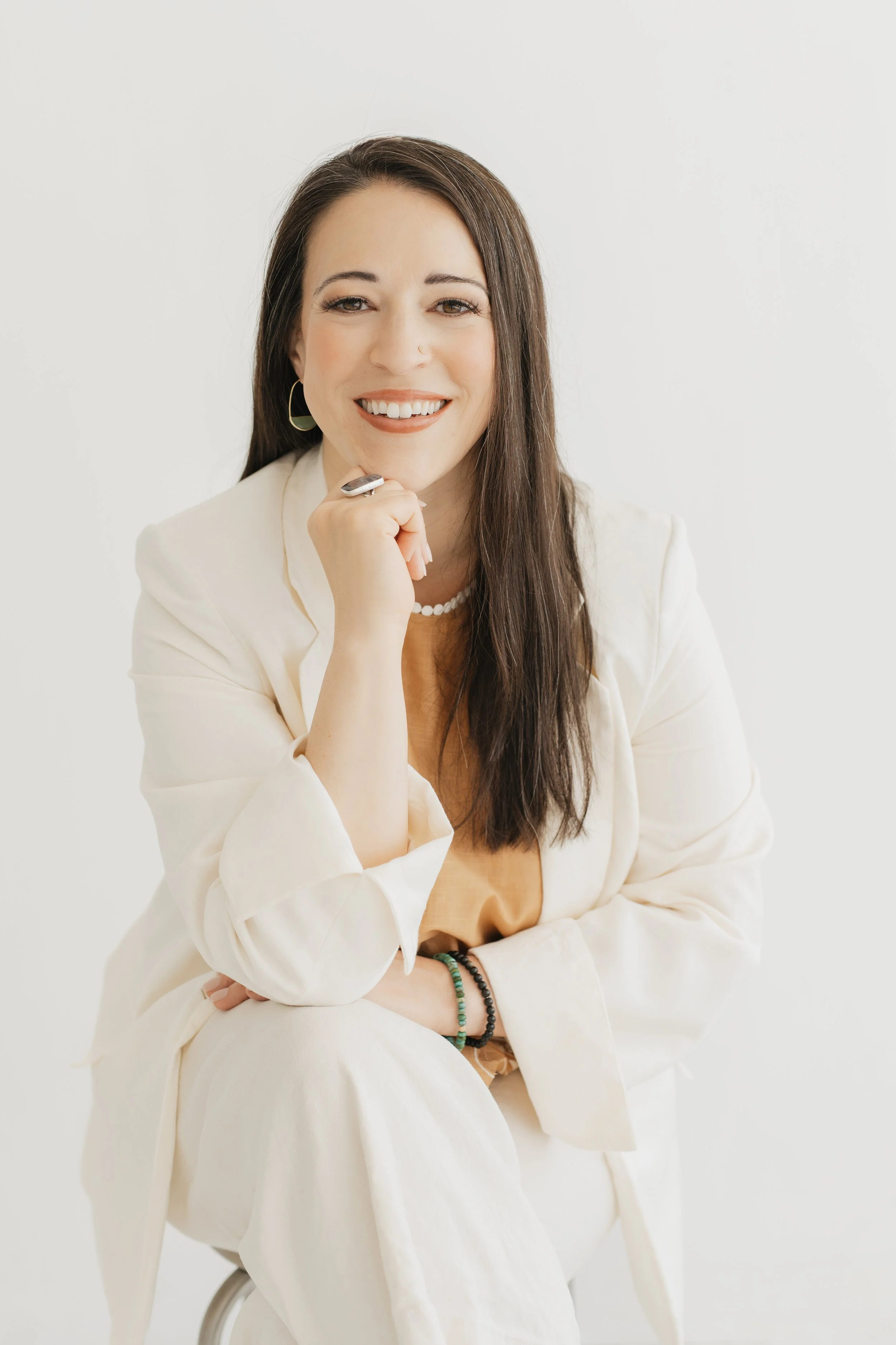 A young woman with long brown hair and light skin smiling while sitting on a chair against a plain white background. She is wearing a white blazer, a peach-colored top, a pearl necklace, hoop earrings, and bracelets, with her left arm resting on her lap and her right hand touching her chin.