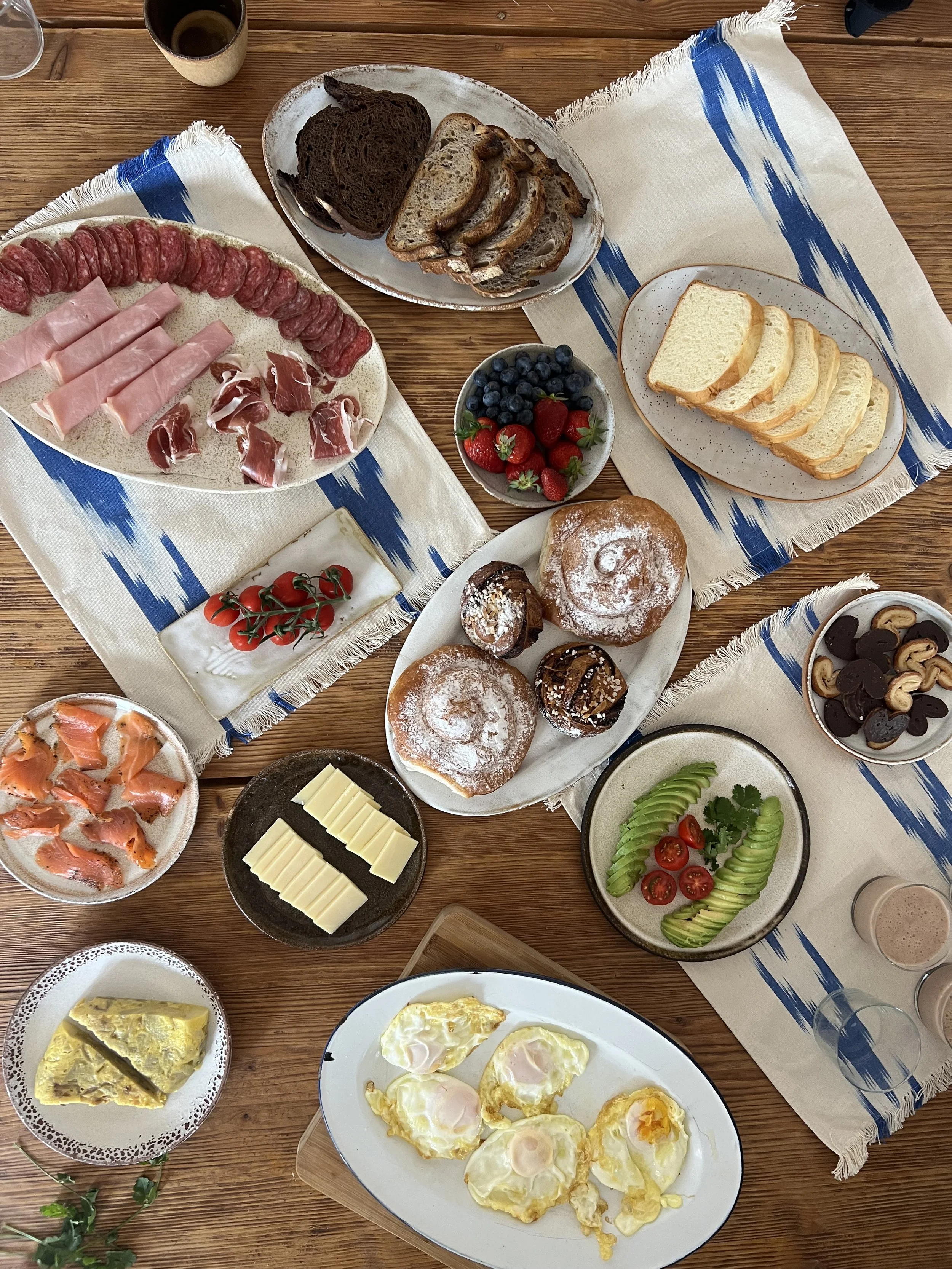 A breakfast spread on a wooden table with various plates of food. There are sliced bread, croissants, and muffins, sliced cured meats like salami, ham, and prosciutto, a bowl of strawberries and blueberries, a plate of sunny side up eggs, a bowl of cherry tomatoes and cucumber slices, and a small dish of white cheese squares. There are also cups of drinks and decorative cloths underneath the plates.