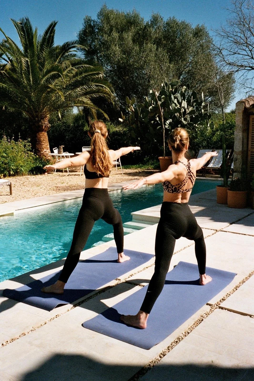 Two women practicing yoga on mats beside a pool outdoors, with trees in the background on a sunny day.