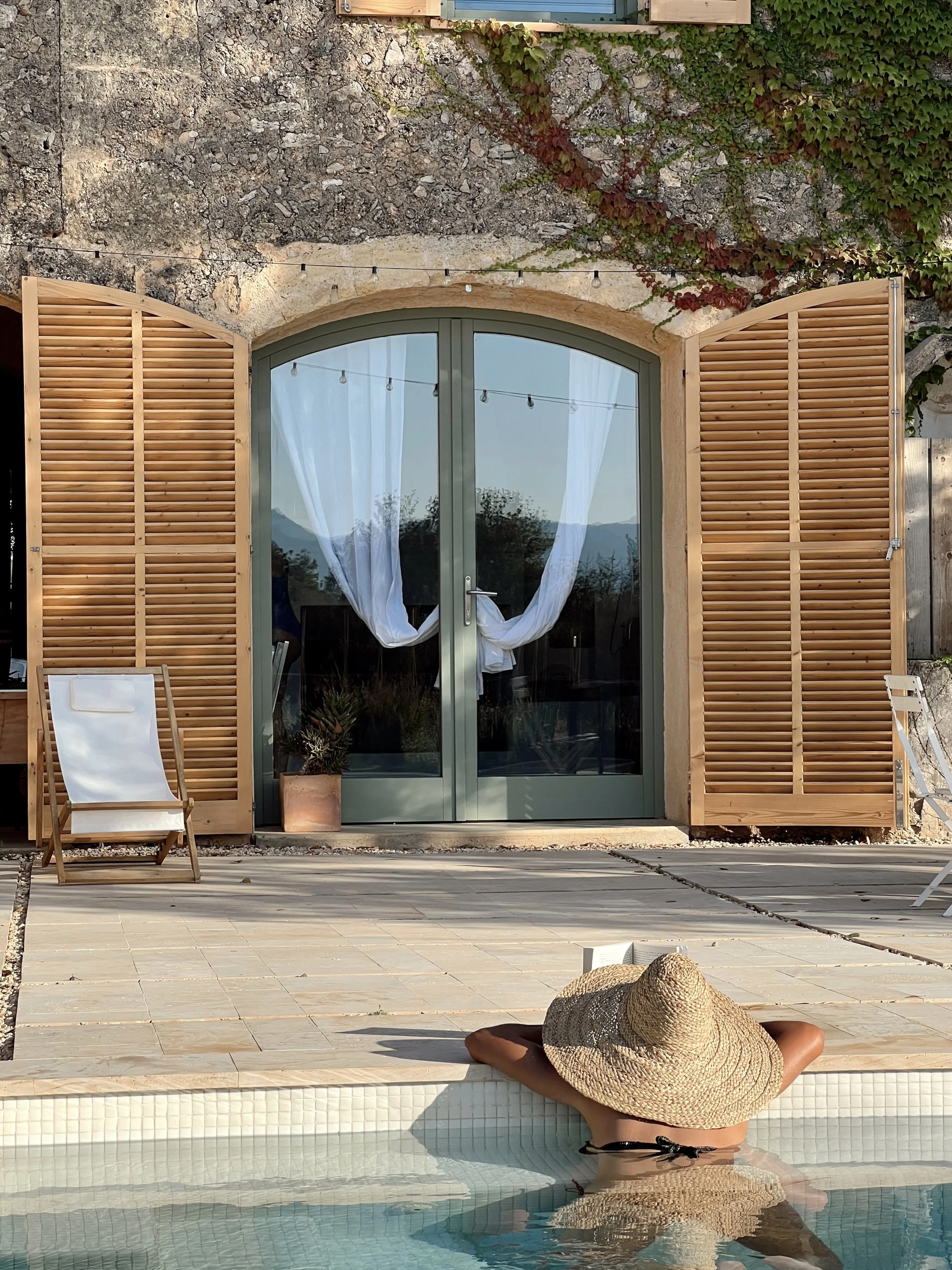 Woman relaxing in a swimming pool wearing a large straw hat and a black swimsuit, with her back to the camera. The pool has a tiled edge, and there is a tan hat on the tiled surface in front of her. Behind her, there is an outdoor patio area with wooden chairs and a potted plant. The background features a stone wall with green and red ivy and a window with blue shutters.