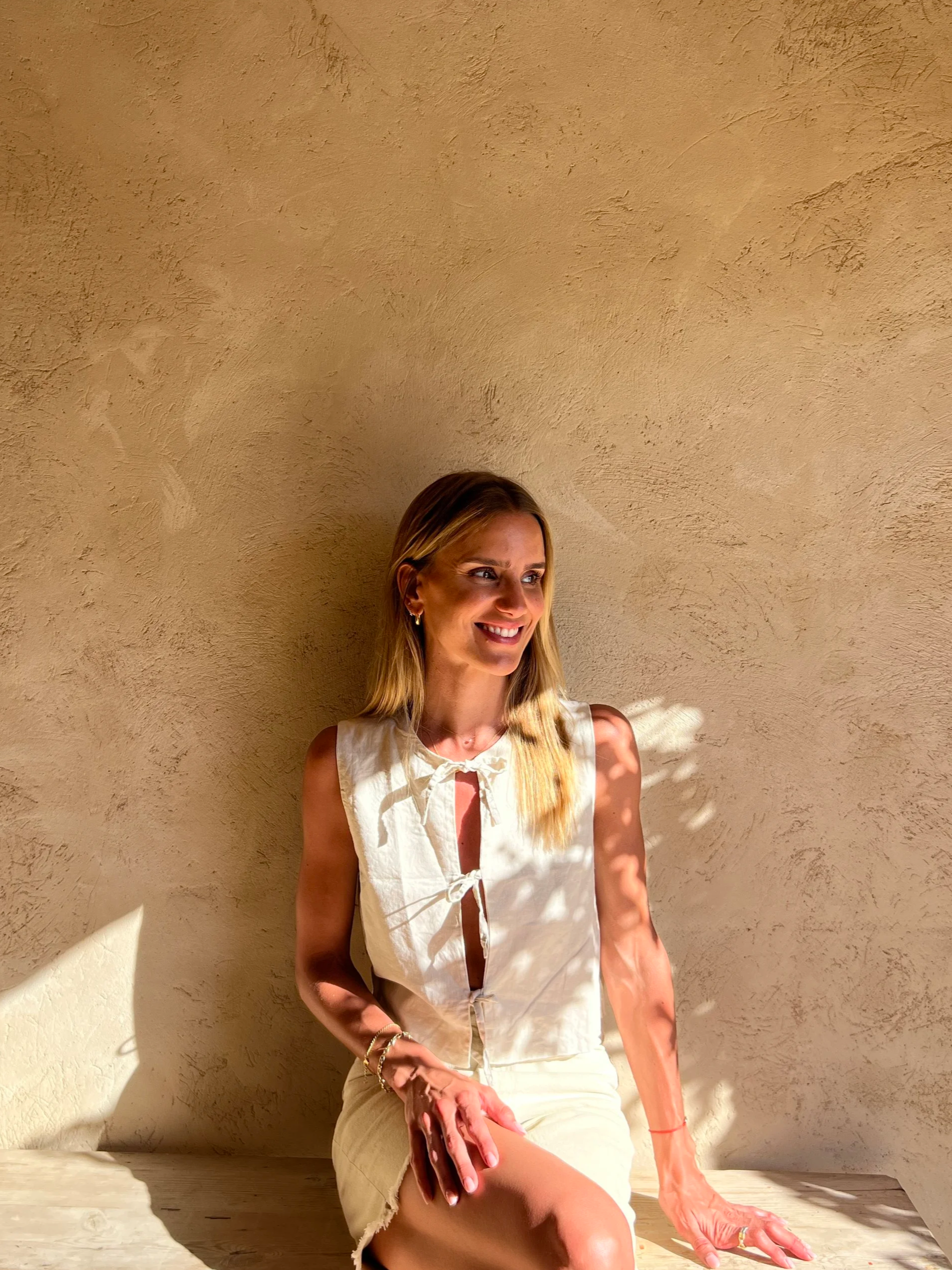 A woman with long blonde hair smiling, wearing a sleeveless white top with a tie in front and a white skirt, sitting against a textured beige wall with sunlight casting shadows.