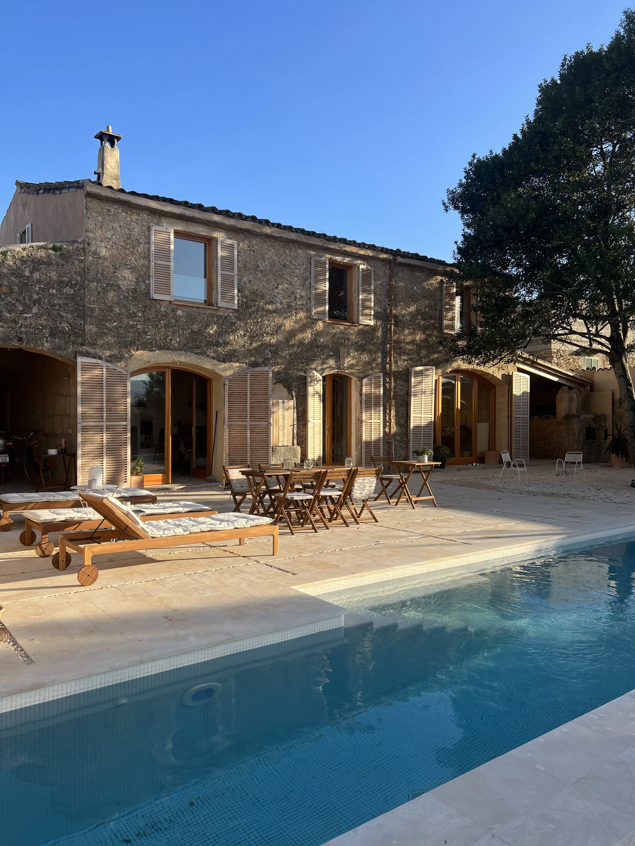 A stone house with wooden shutters and a swimming pool with lounge chairs and tables in the outdoor patio area, under a clear blue sky.