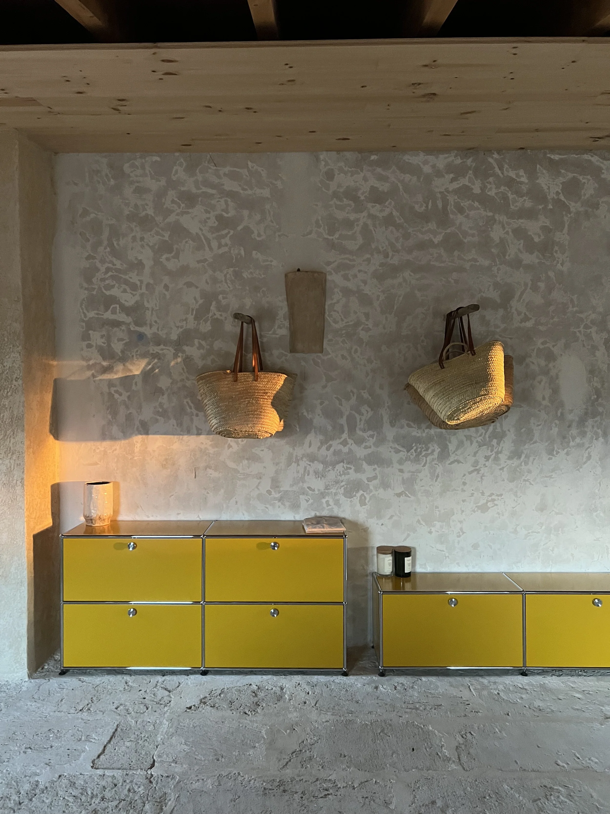 Yellow cabinet with eight drawers and metal legs, alongside a smaller table with two jars. Above the cabinet, two wicker wall-mounted baskets with leather straps are hanging on a textured wall. A small wooden shelf and a candle holder are on the left side, with a glass candle holder on the table.