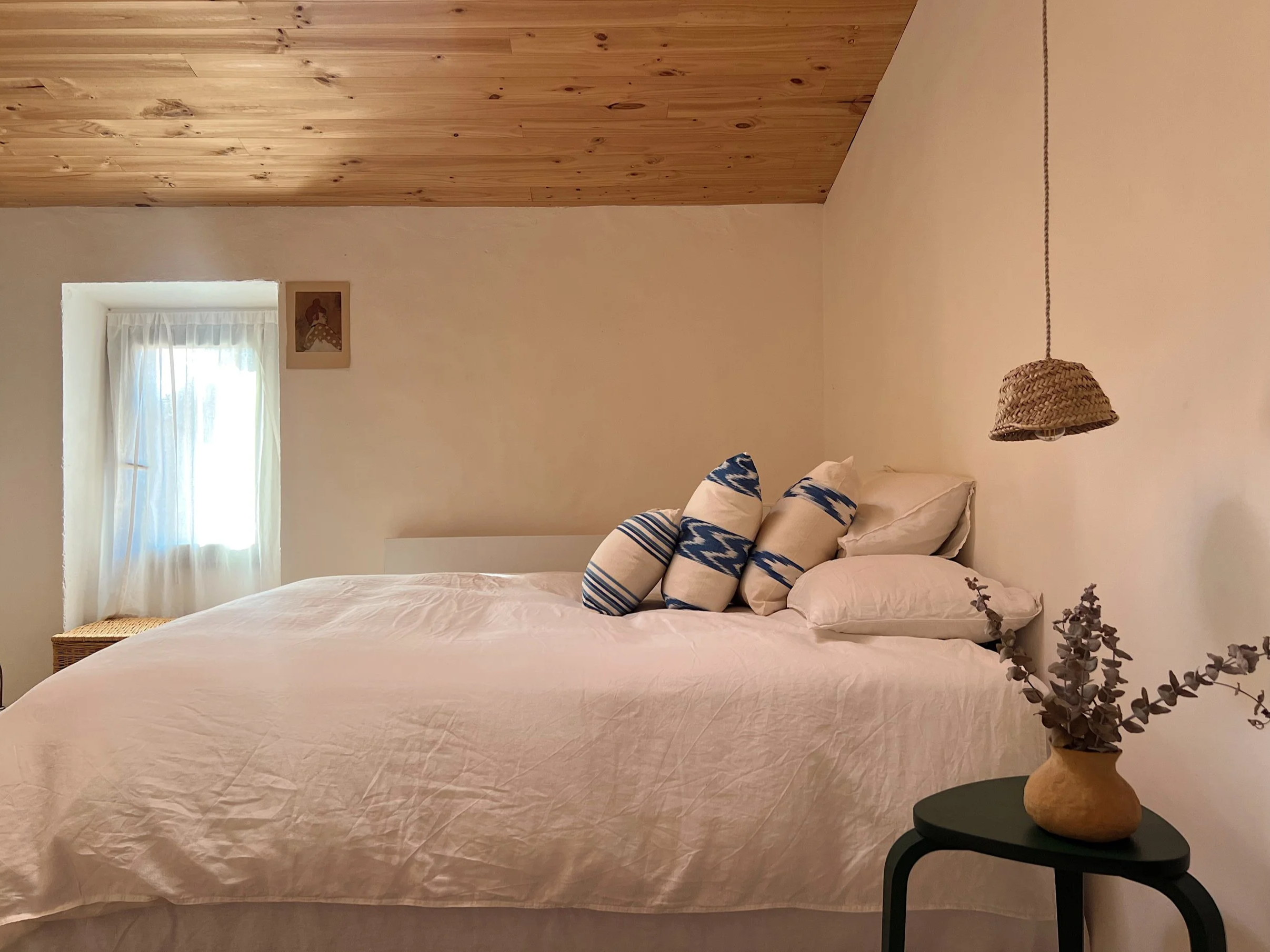 A bedroom with a bed covered in a light pink bedspread, adorned with striped pillows in shades of blue and white. To the right, there is a black side table holding a beige woven lampshade and a potted plant with purple leaves. The wall behind the bed is painted white and has a small window with sheer white curtains, allowing natural light into the room. There is a framed picture hanging on the wall above the window. The ceiling features wood paneling.