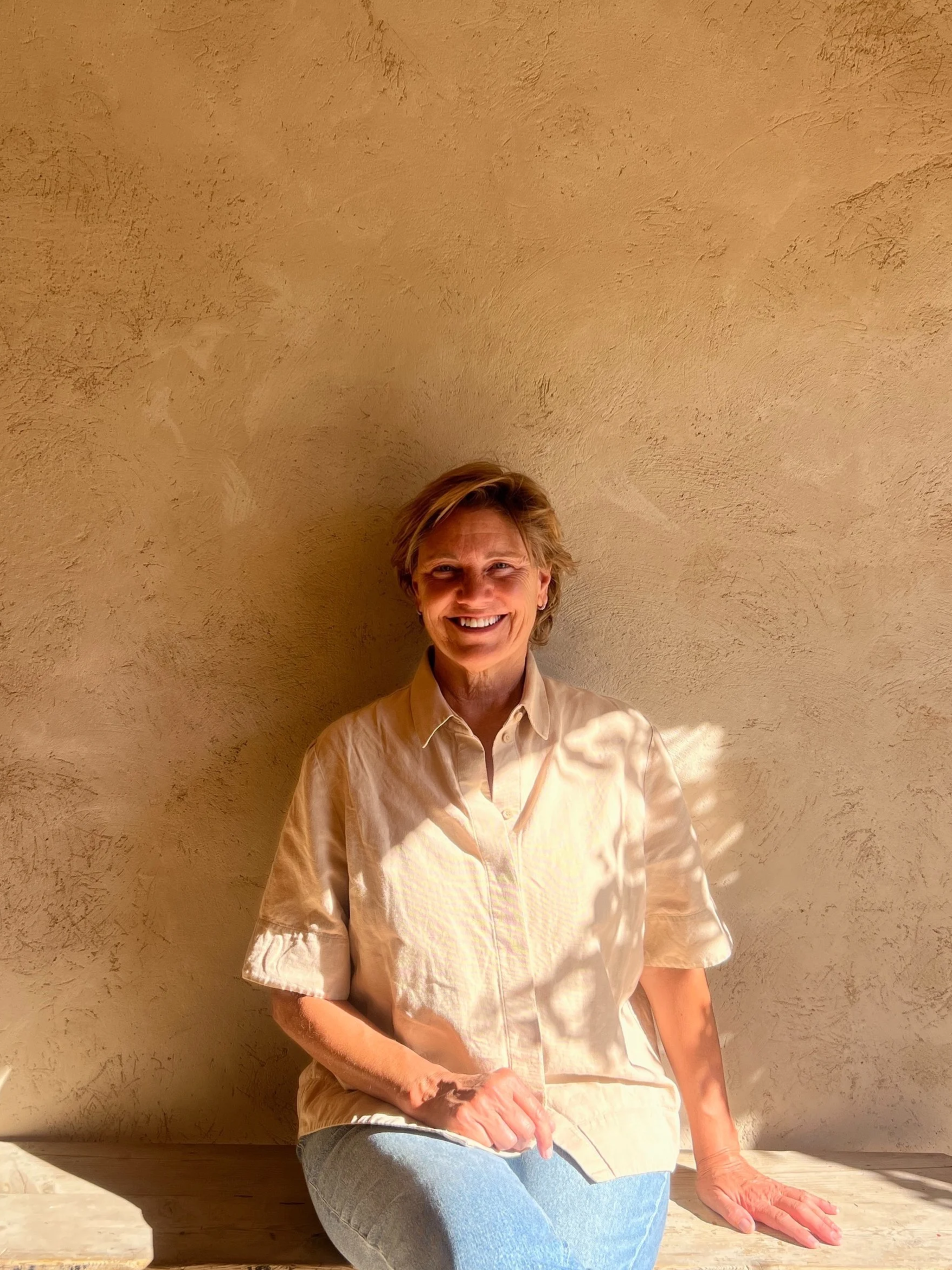 Person with short, light brown hair smiling, wearing a beige button-up shirt and blue jeans, sitting on a wooden surface with a textured beige wall behind them.