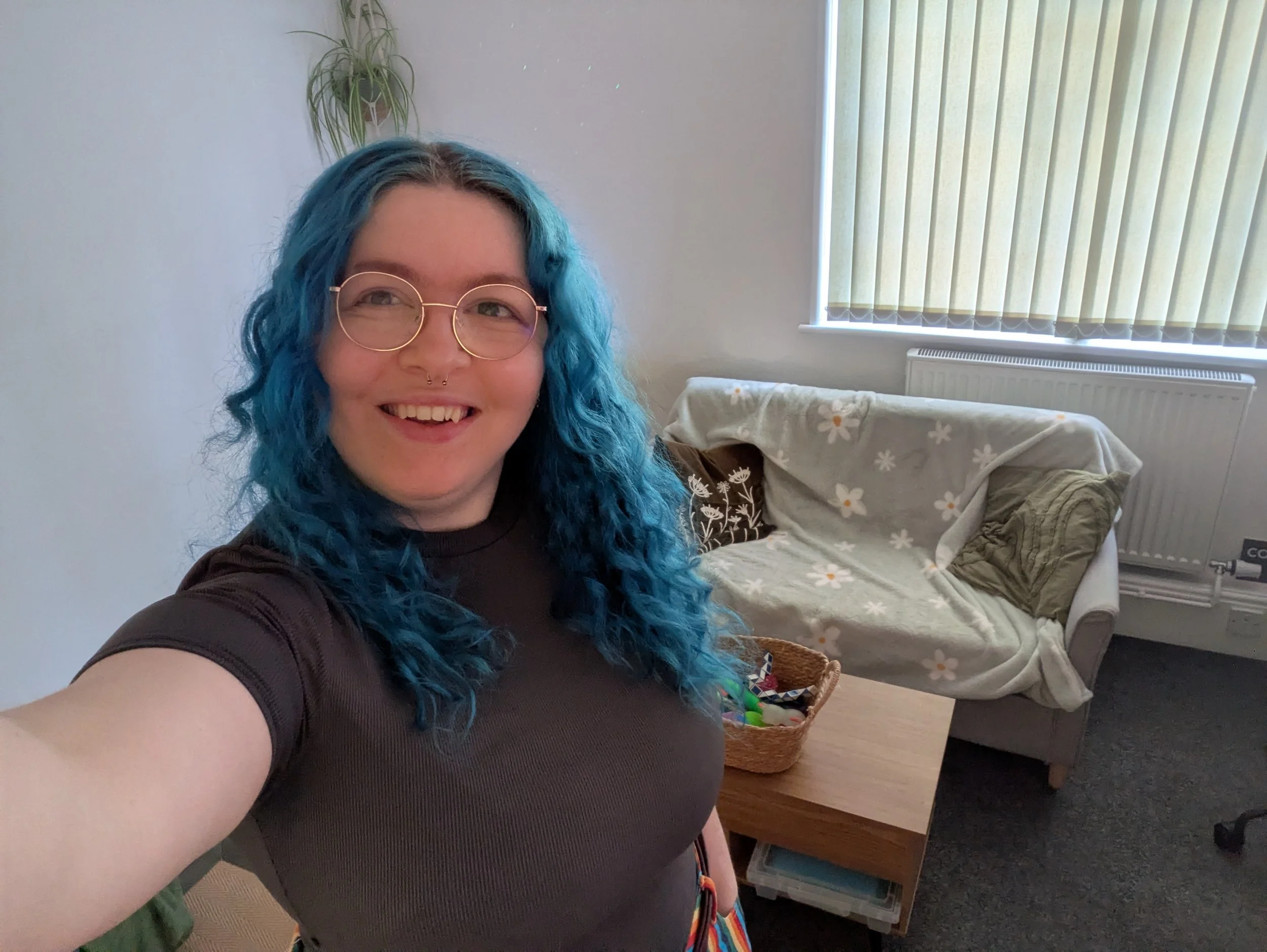 A therapist with curly blue hair and glasses smiling in a room with a sofa covered with a daisy-patterned blanket, a basket of toys, and vertical blinds on the window.