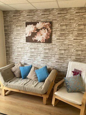 Therapy room with a beige couch with two gray pillows and three blue pillows, and a white armchair with a floral pillow, against a stone accent wall with a painting of pink flowers.