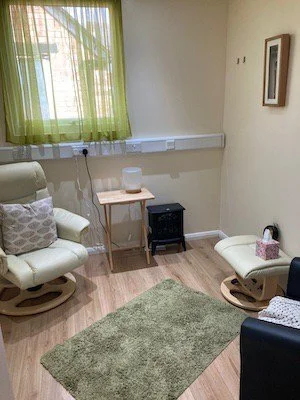 A cozy room with a green curtain, two white rocking chairs, a beige armchair, a small table, and a green rug on light hardwood flooring.