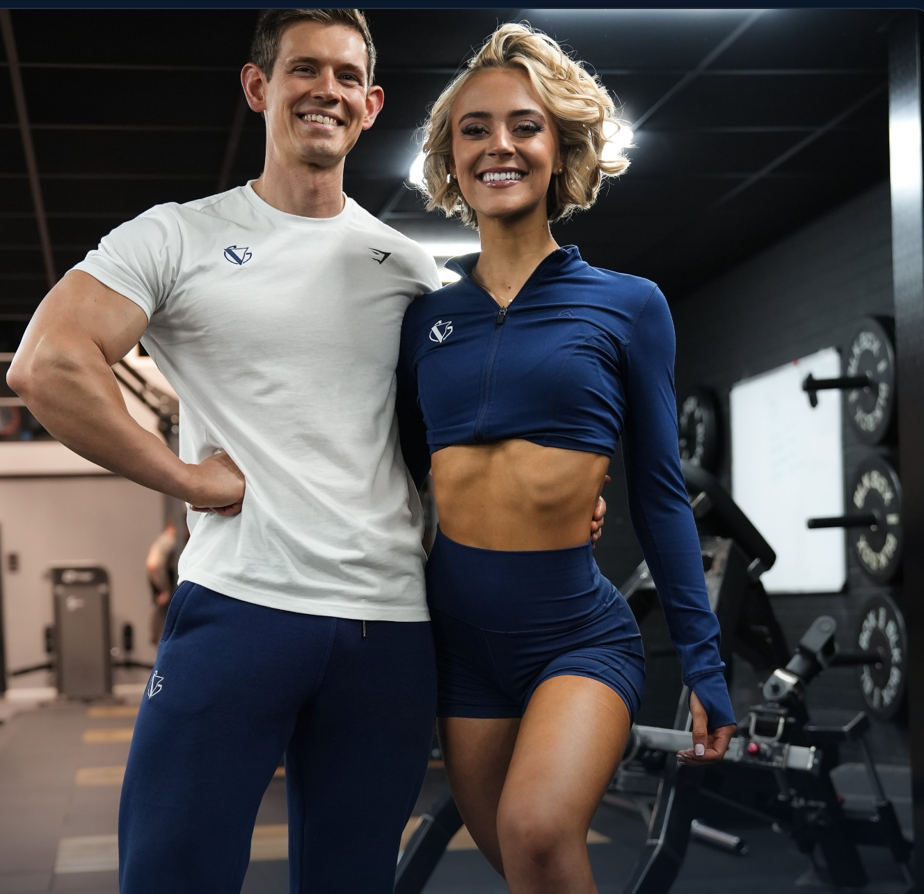 A man and woman at a gym, both smiling and wearing workout clothes. The man has short dark hair and is in a white t-shirt and blue sweatpants, while the woman has short blonde hair and is in a blue sports bra and shorts. The background shows gym equipment and weights.