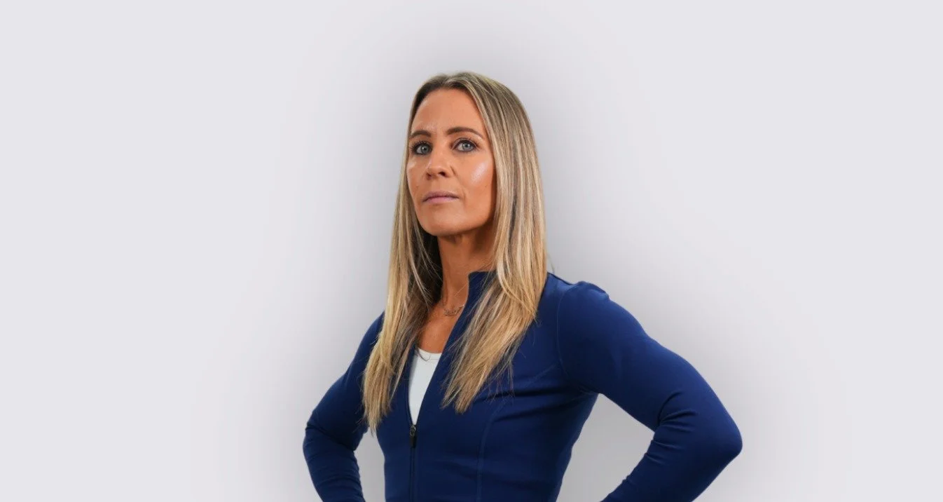 A woman with long blonde hair wearing a blue zip-up jacket and standing with her hands on her hips against a plain light gray background.