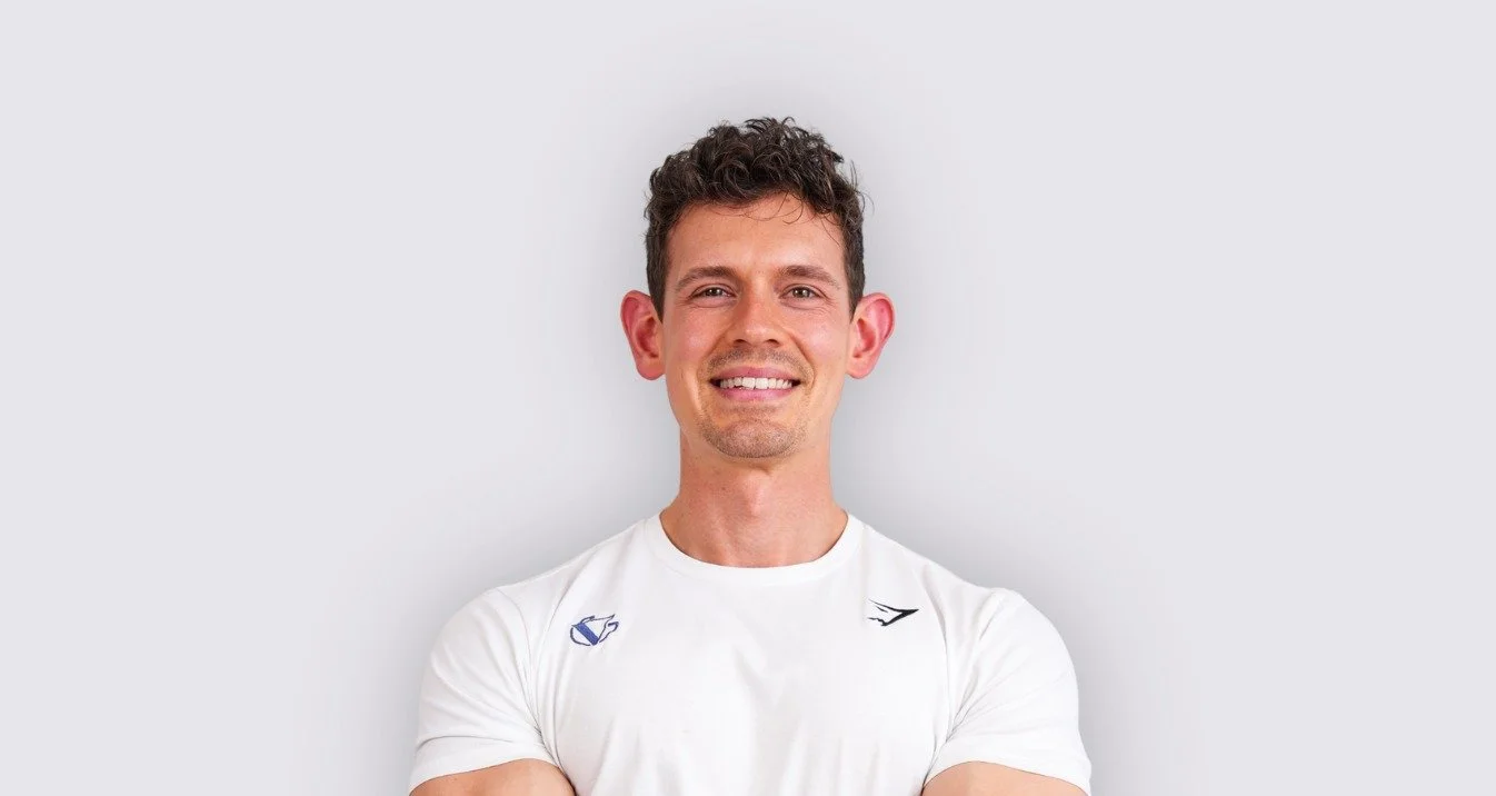 A young man with curly brown hair smiling with arms folded, wearing a white t-shirt, against a plain light gray background.