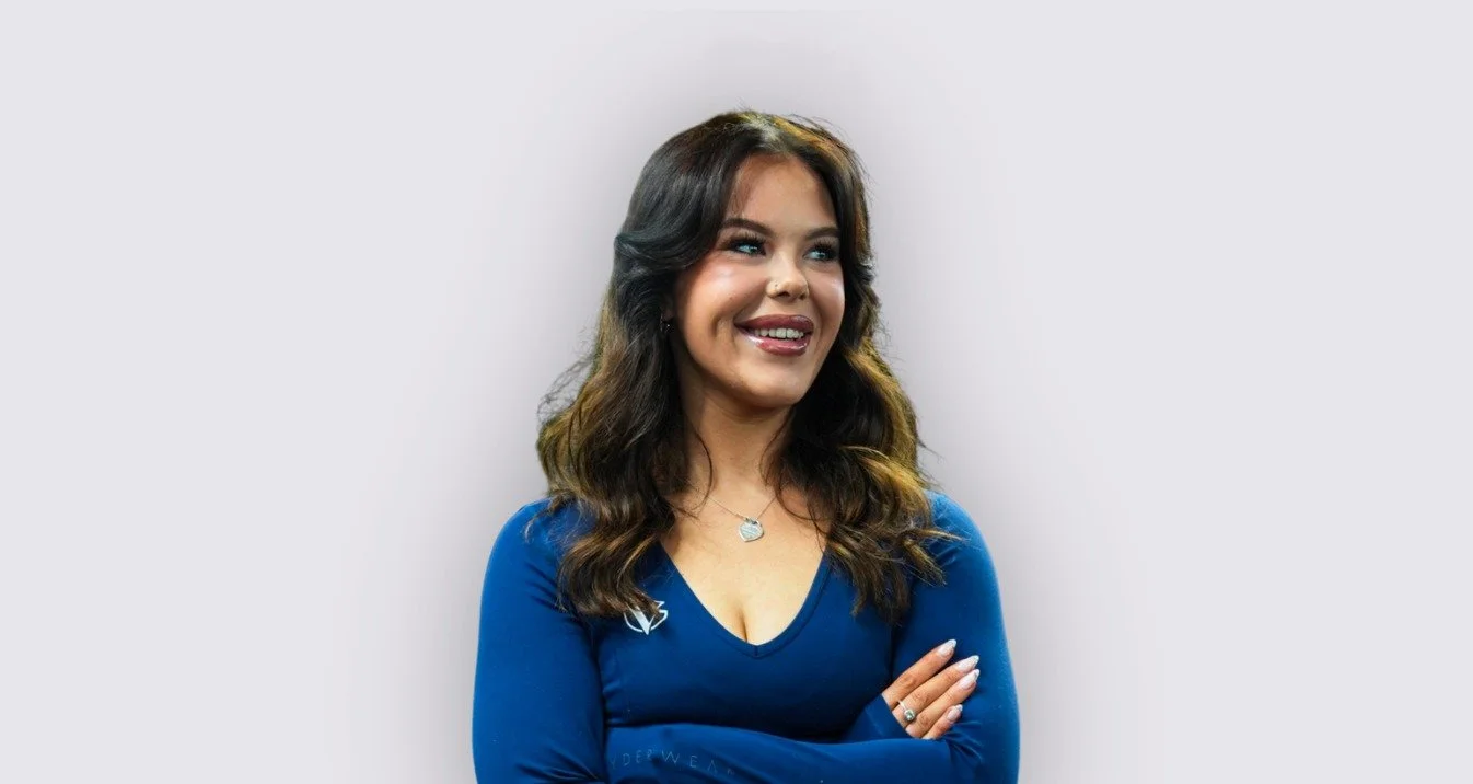 A woman with long, wavy dark hair is smiling and looking to the side. She is wearing a blue long-sleeve top and has a necklace and rings on her fingers. She is standing against a plain light grey background.