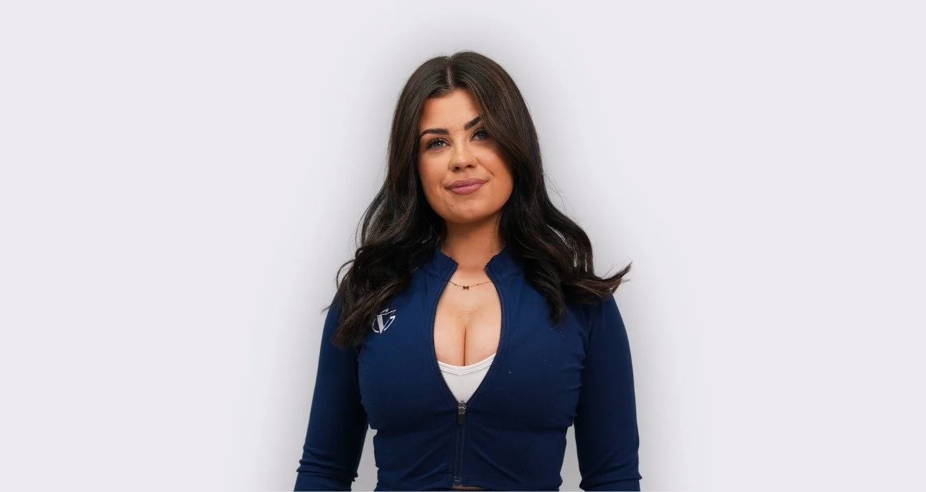 A woman with long dark hair wearing a navy blue zip-up jacket with a white top underneath, standing against a plain white wall.