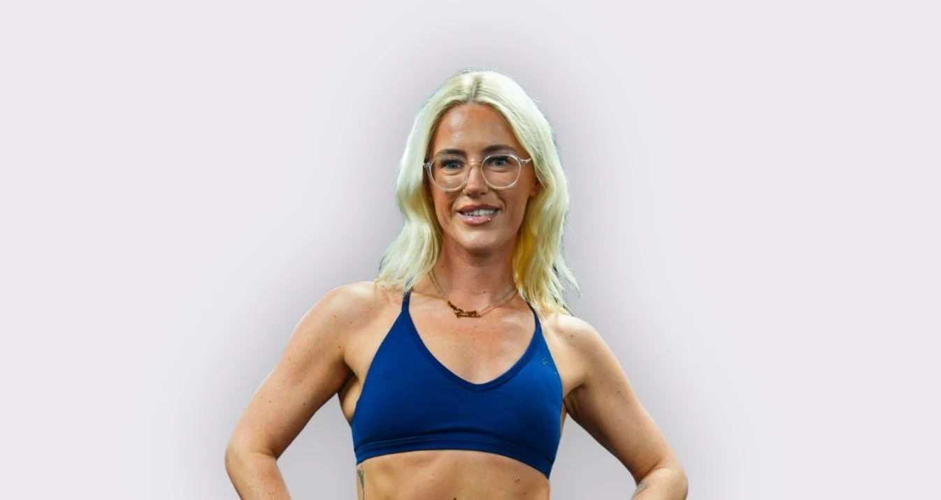 Woman with blonde hair, glasses, and a blue sports bra smiling, standing with her hands on her hips against a plain light background.