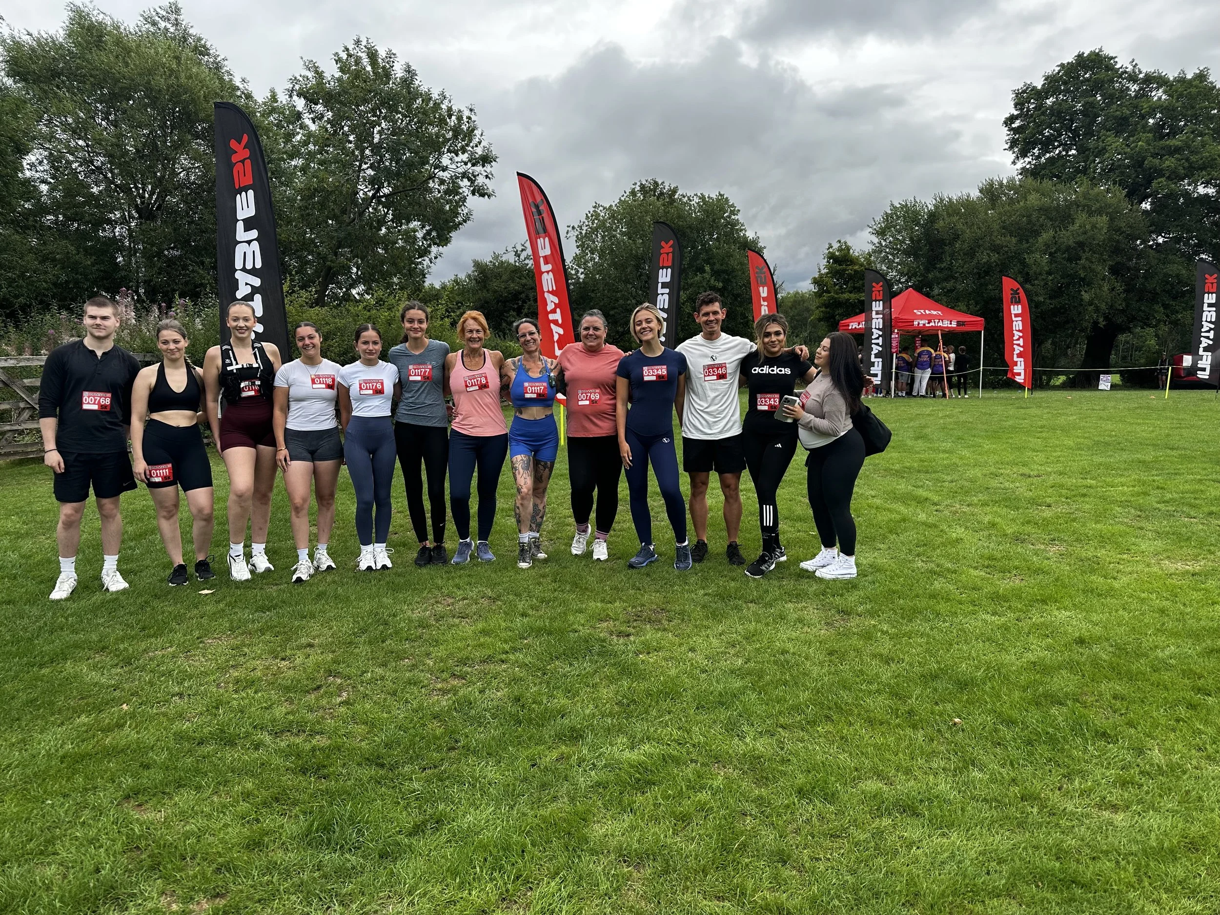 A group of twelve people, consisting of a mix of men and women, standing on a grassy field during a cloudy day at a running event. They are wearing athletic clothing and have race bibs pinned to their shirts, with a few smiling at the camera. In the background, there are tall black and red flags and a red tent with more participants nearby.
